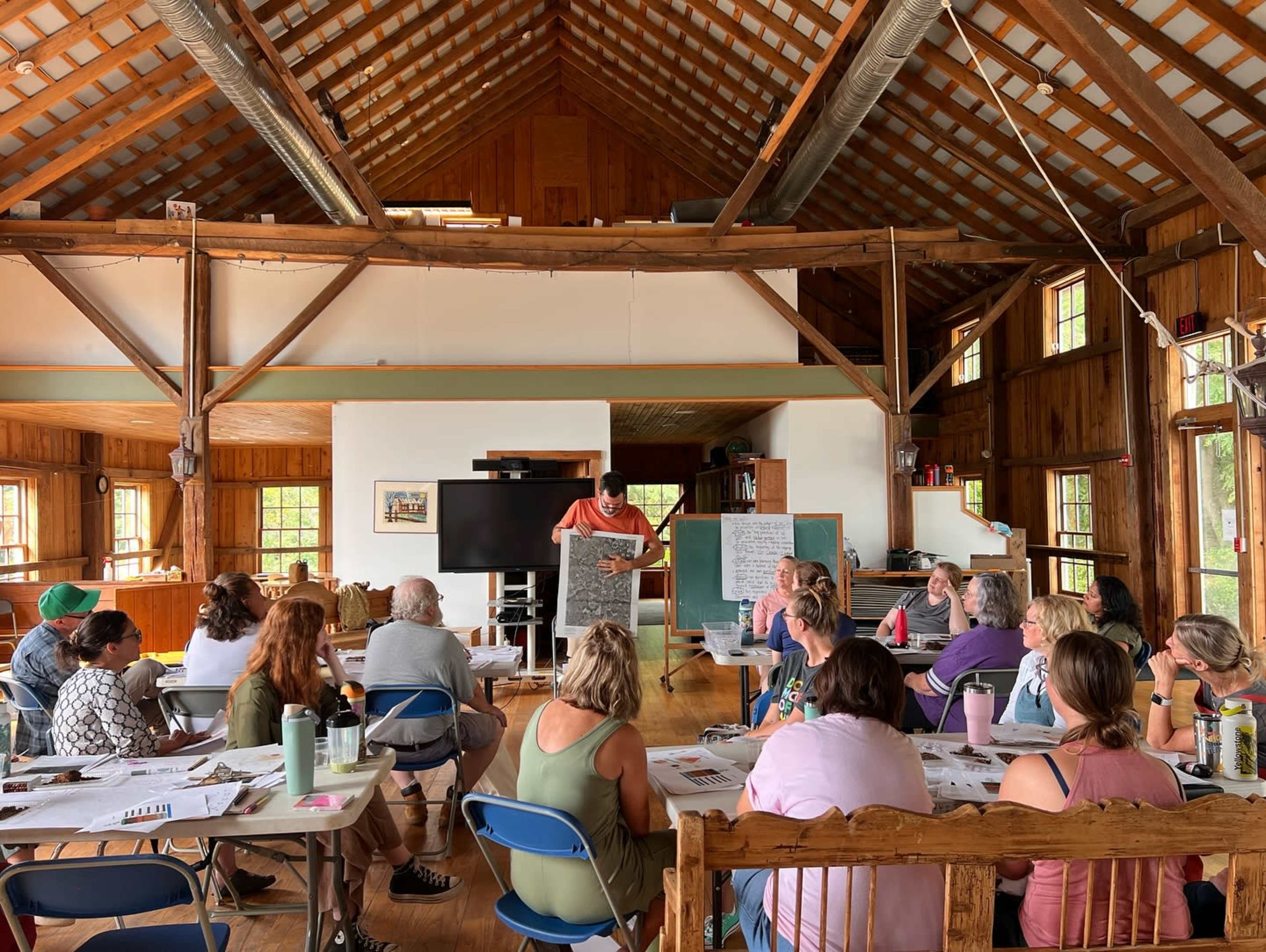 A group of individuals is seated in a rustic wooden building with a high ceiling, listening to a speaker presenting at the front of the room.