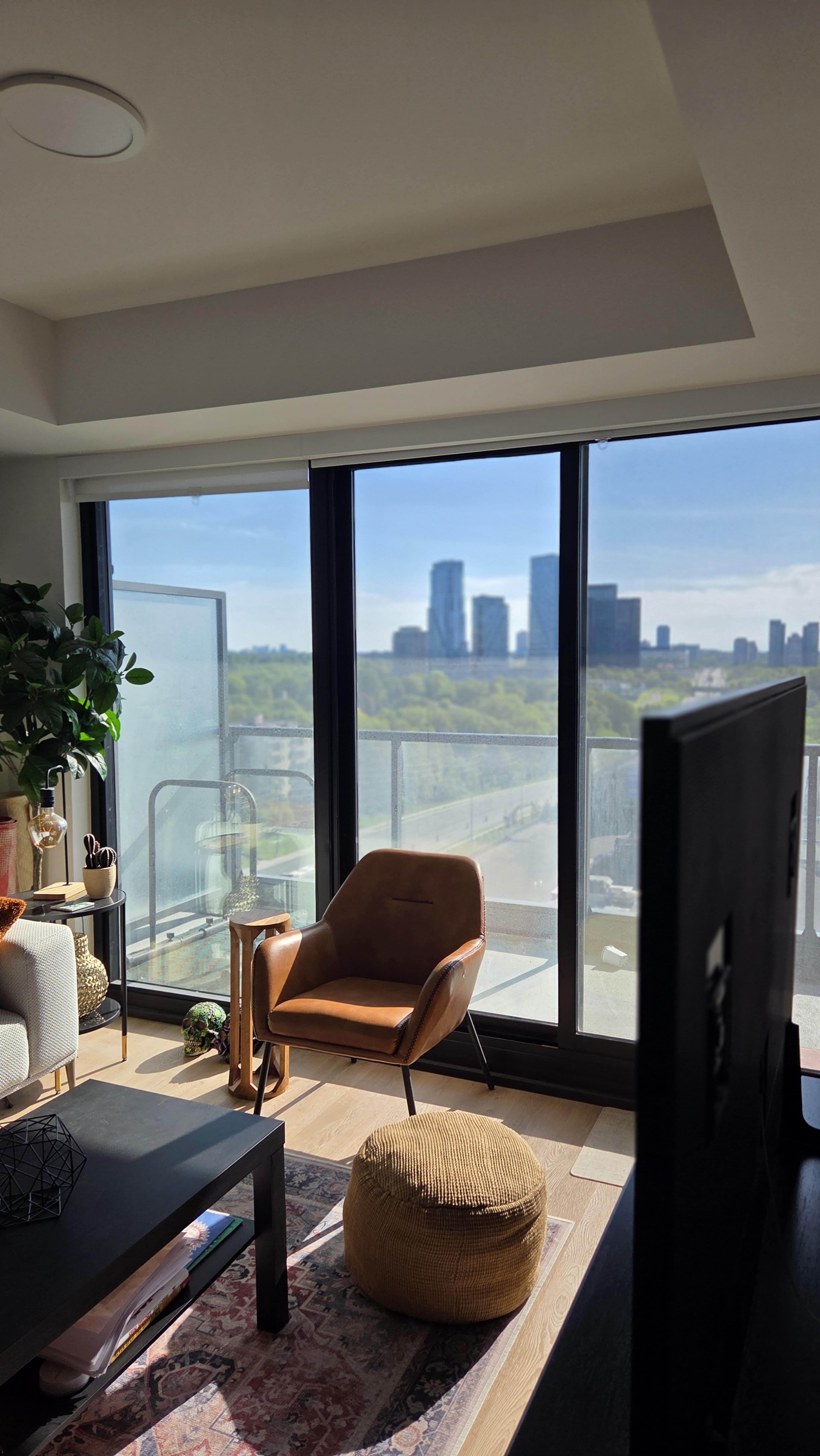 A modern living room features a brown armchair and a round ottoman, with large windows offering a view of a city skyline and green trees outside.