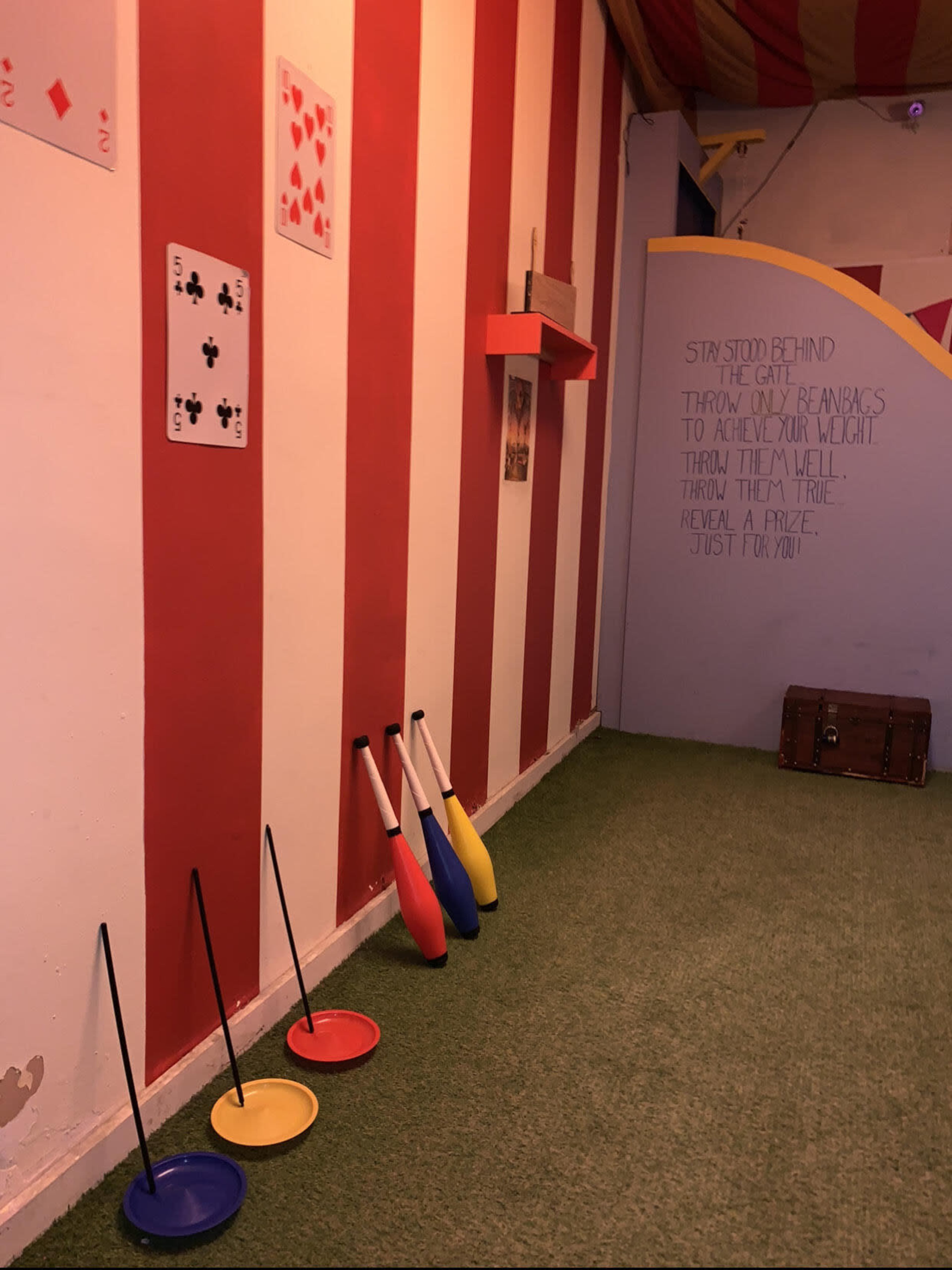A brightly colored game room with red and white striped walls, featuring large playing cards and several juggling pins arranged on the floor.