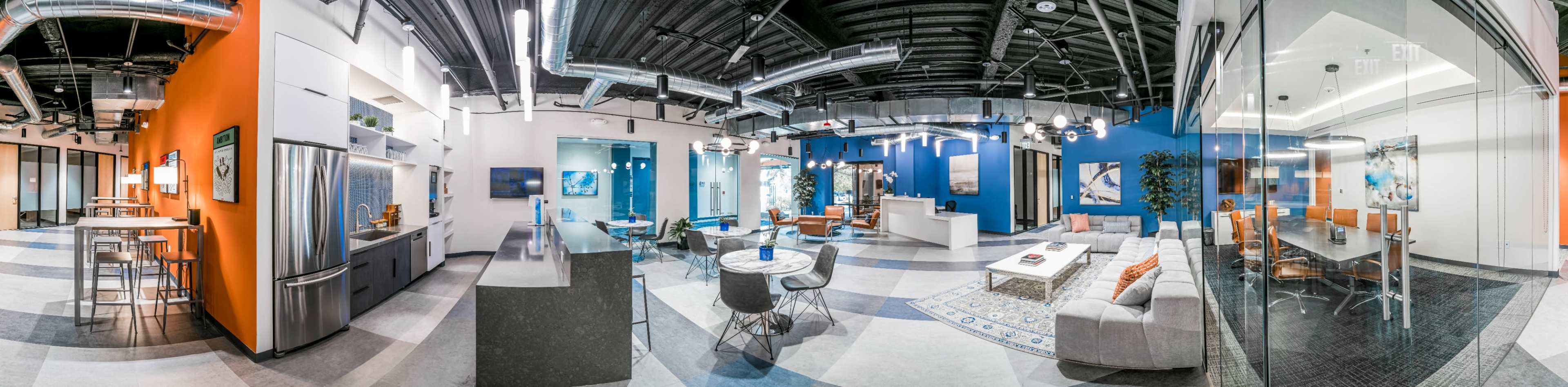 A modern office space featuring open seating areas, a kitchen with a bar, and glass-walled meeting rooms.