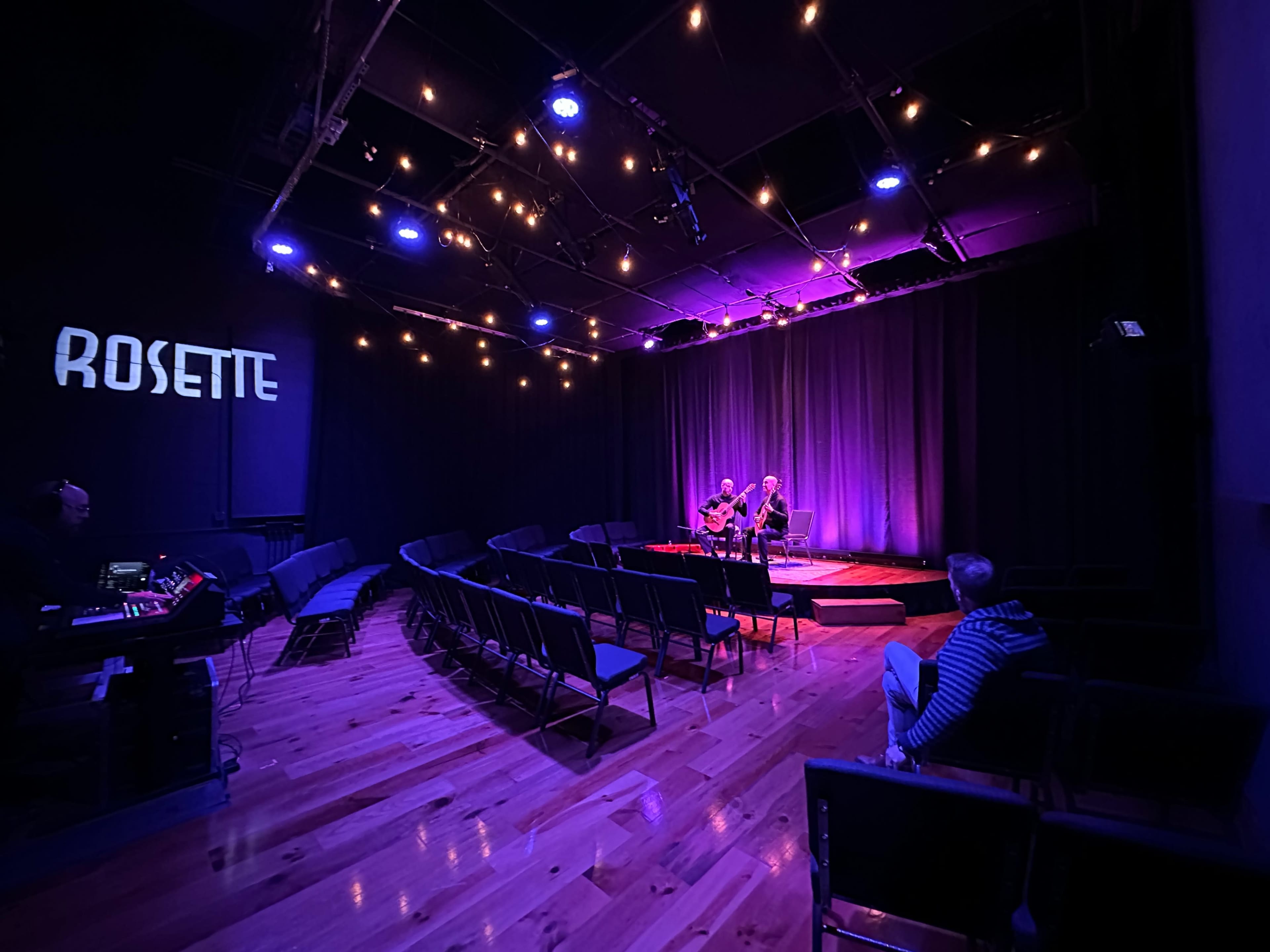 The image shows a dimly lit performance space with empty chairs facing a small stage where two musicians play, illuminated by purple and blue lights.