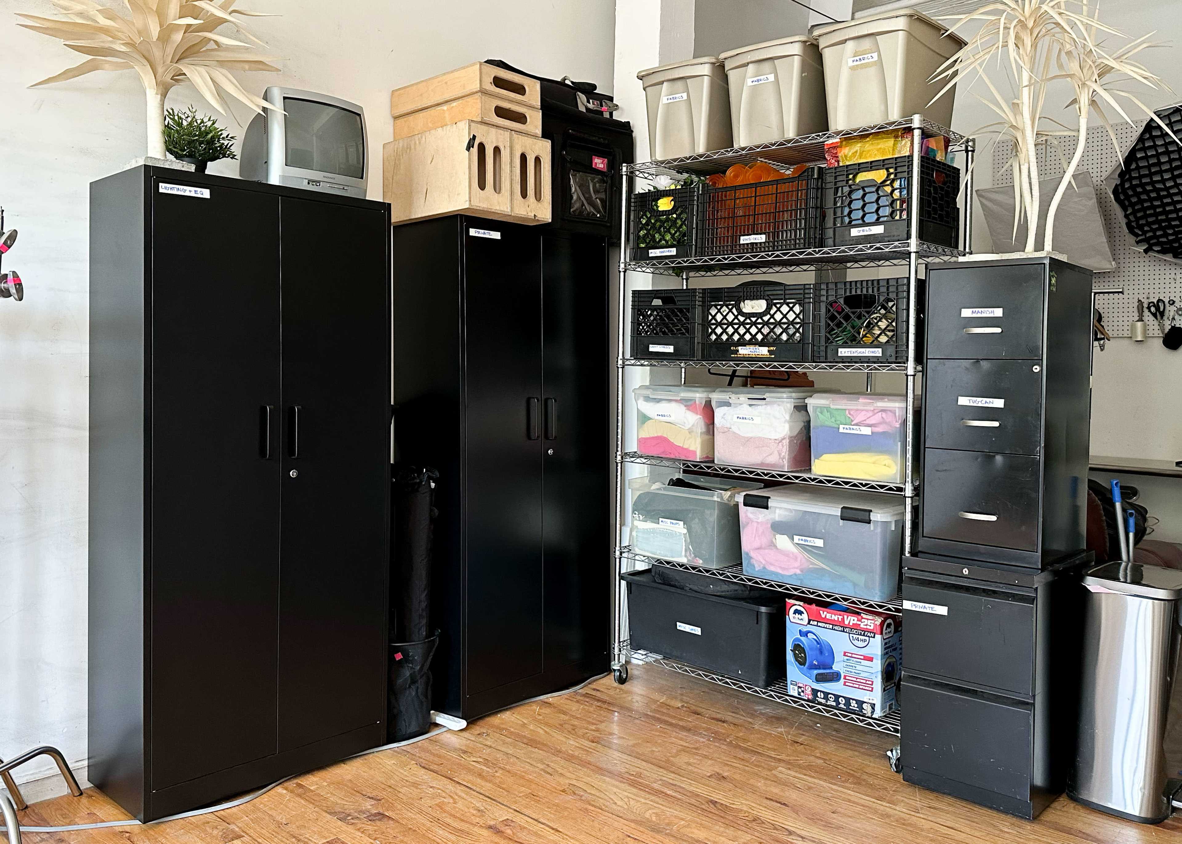 A storage area featuring two black cabinets, several clear bins filled with colorful items, and a variety of labeled containers on metal shelving.
