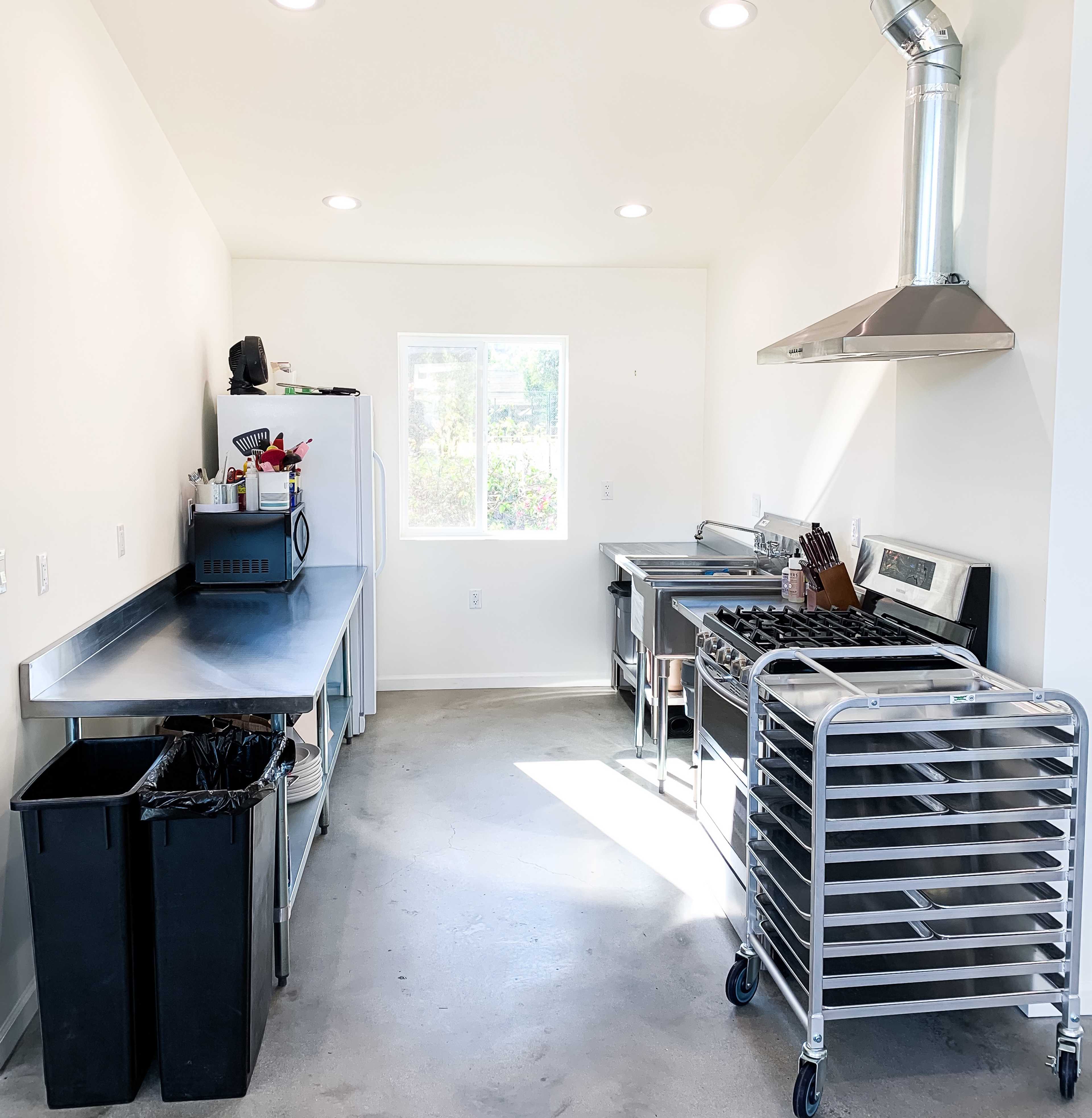 A modern kitchen with stainless steel countertops, a stove, and a rack beside two black trash bins.