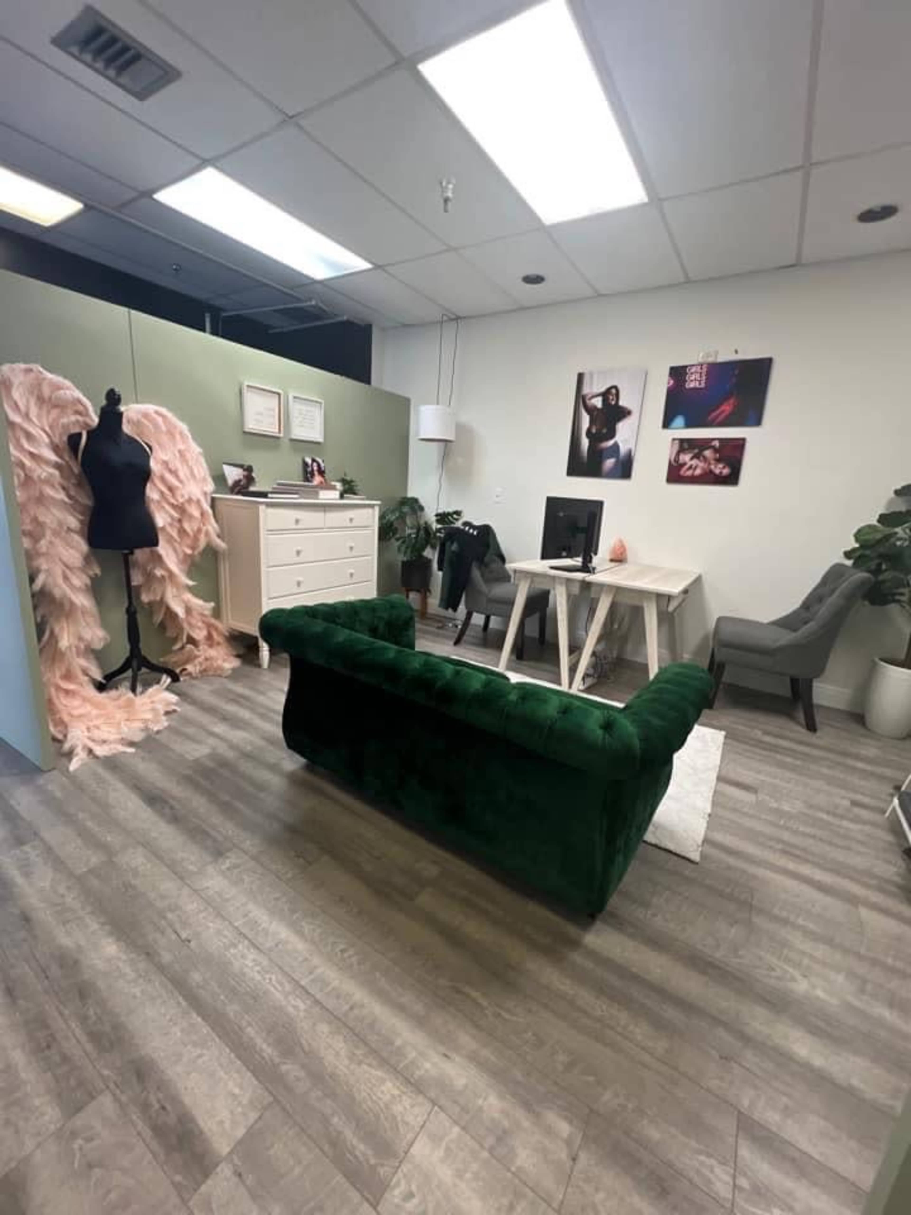 A well-lit office space featuring a green velvet couch, a white desk with a computer, a mannequin dressed in pink fabric, and various plants.