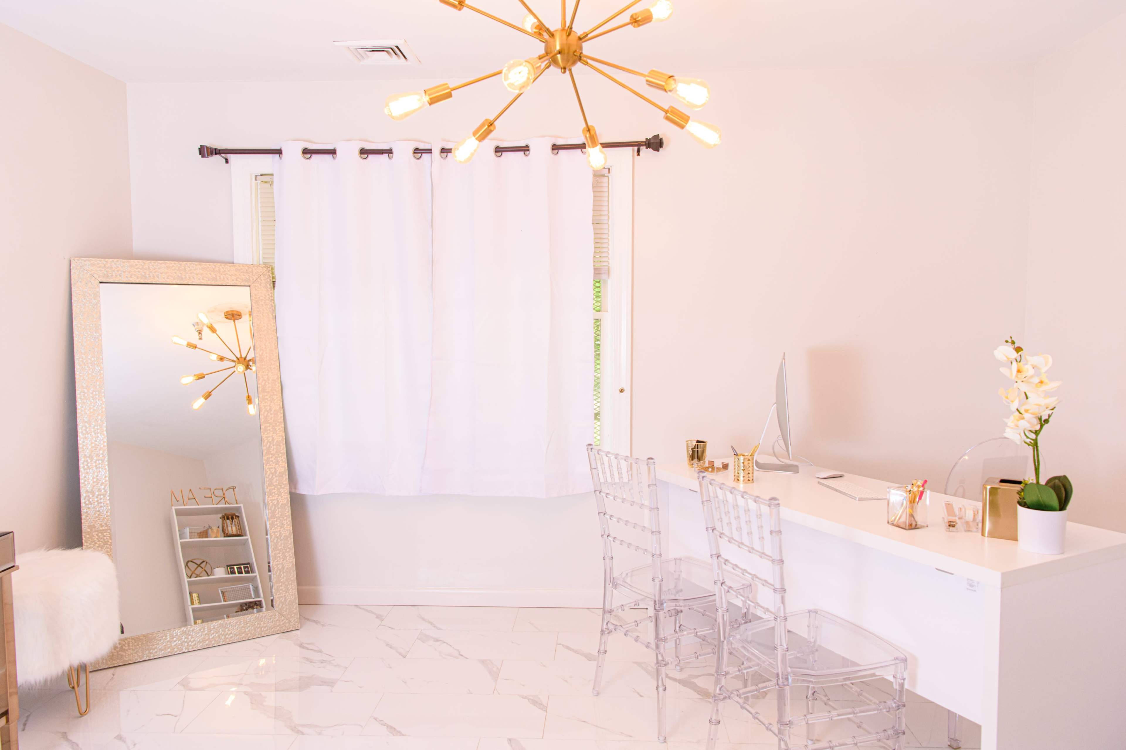 A modern workspace features a white desk with two clear chairs, a large mirror, and a pendant light, all set against a bright, minimalist backdrop.