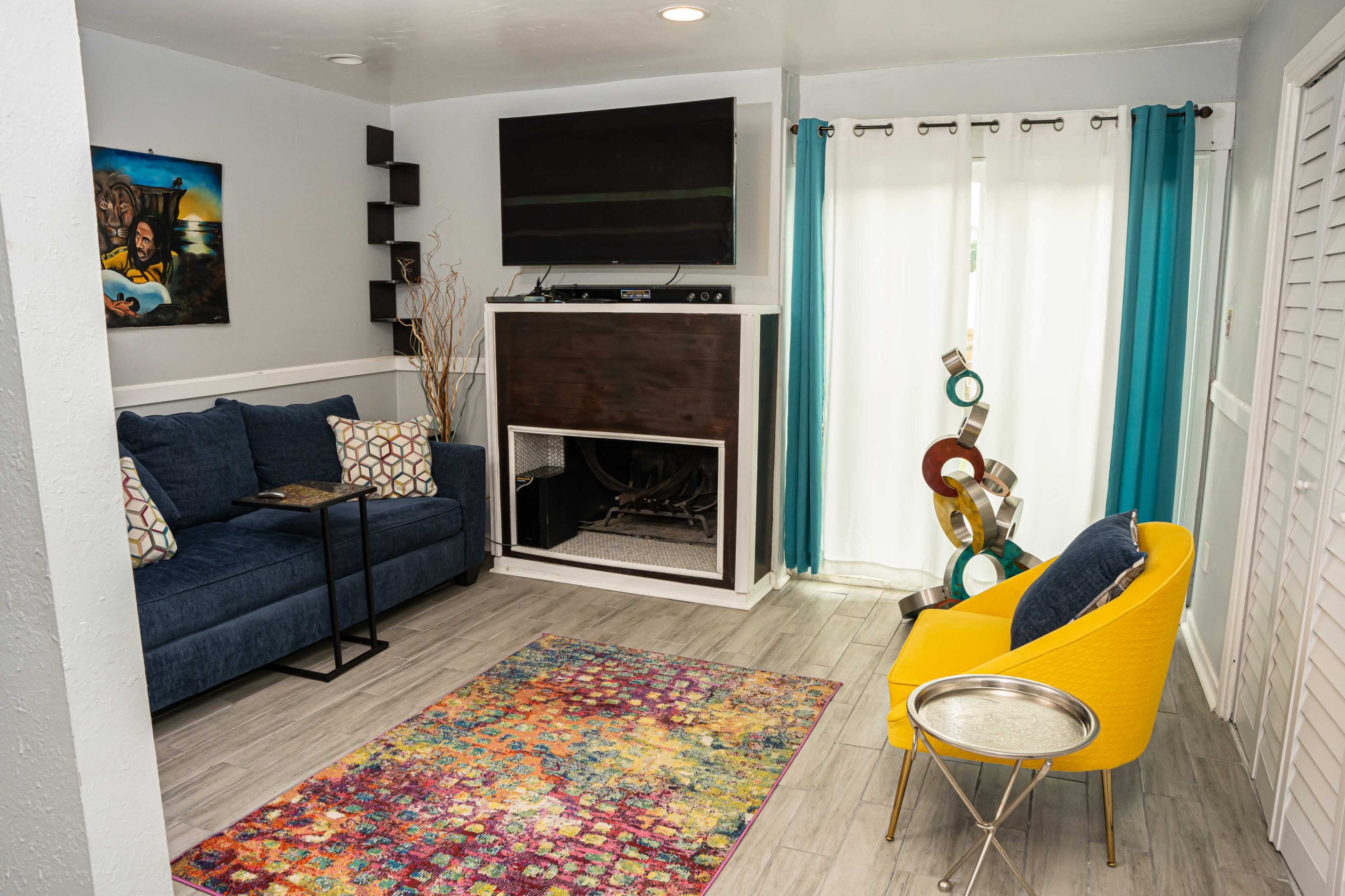 A living room with a blue sofa, a yellow chair, a multicolored area rug, and a fireplace beneath a mounted television, with large windows dressed in sheer curtains.