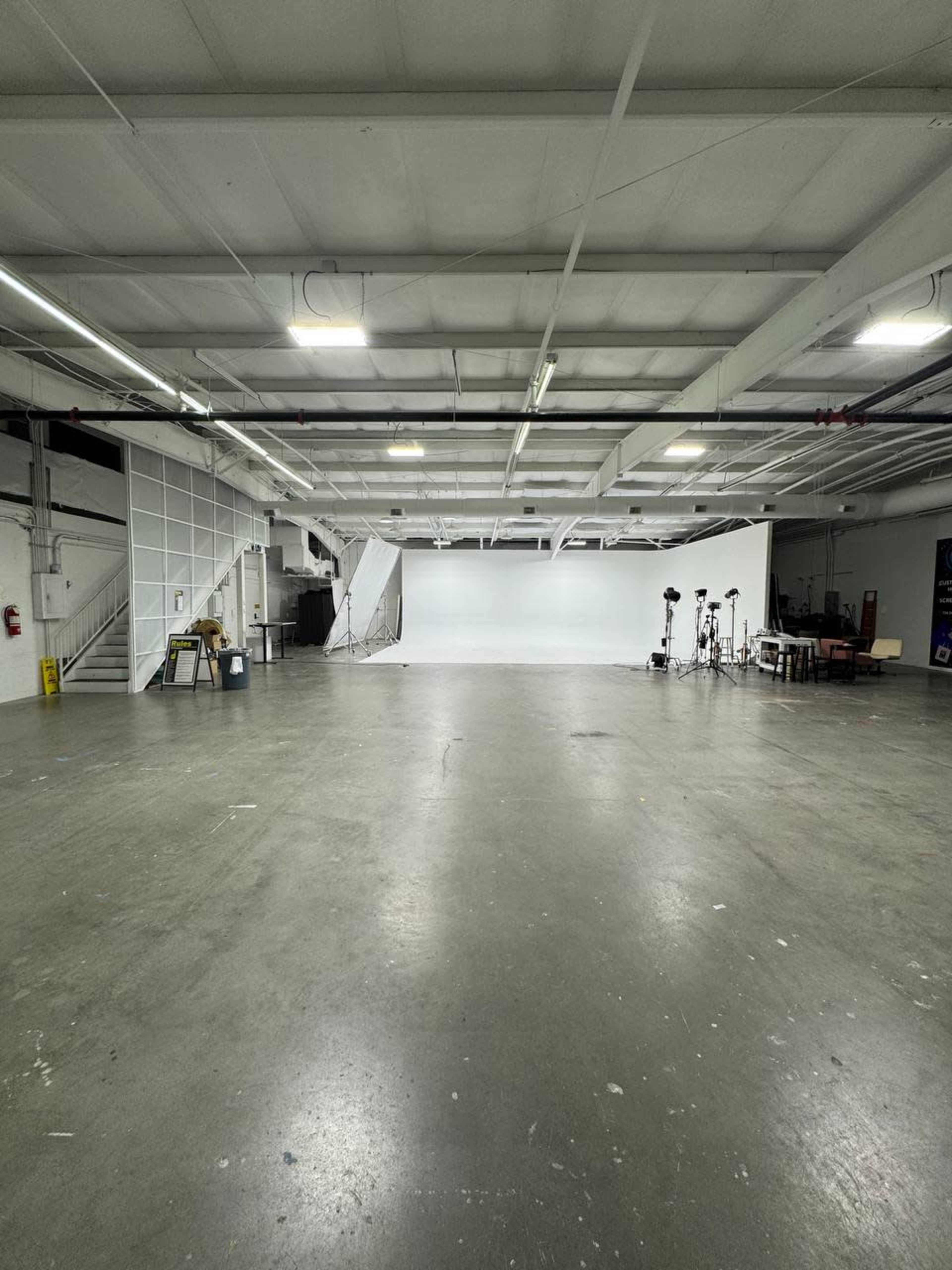 The image shows a large, empty studio space with a white backdrop, various lighting equipment, and a few pieces of furniture.