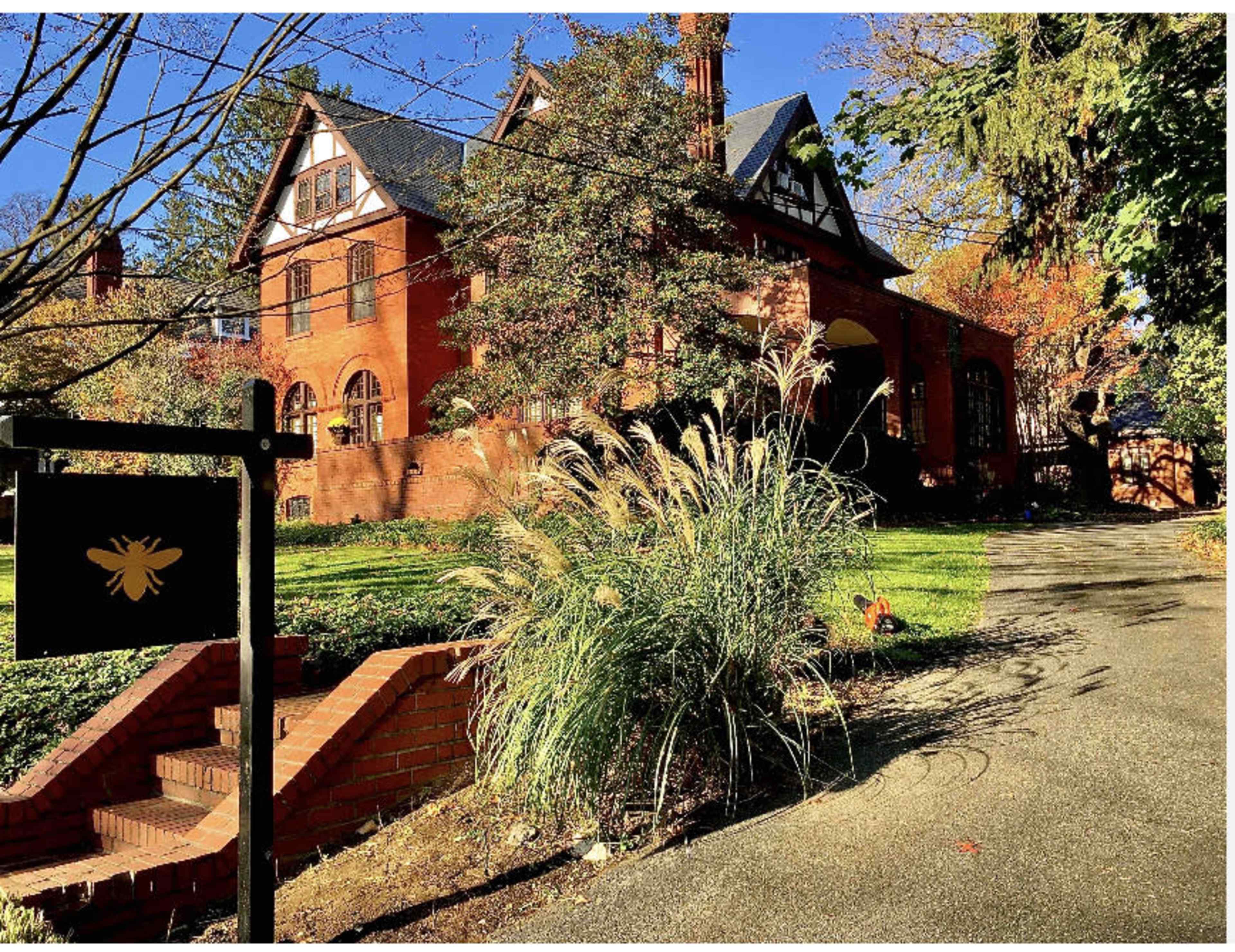 A red brick house with a gabled roof is surrounded by lush greenery and autumn foliage along a winding pathway.