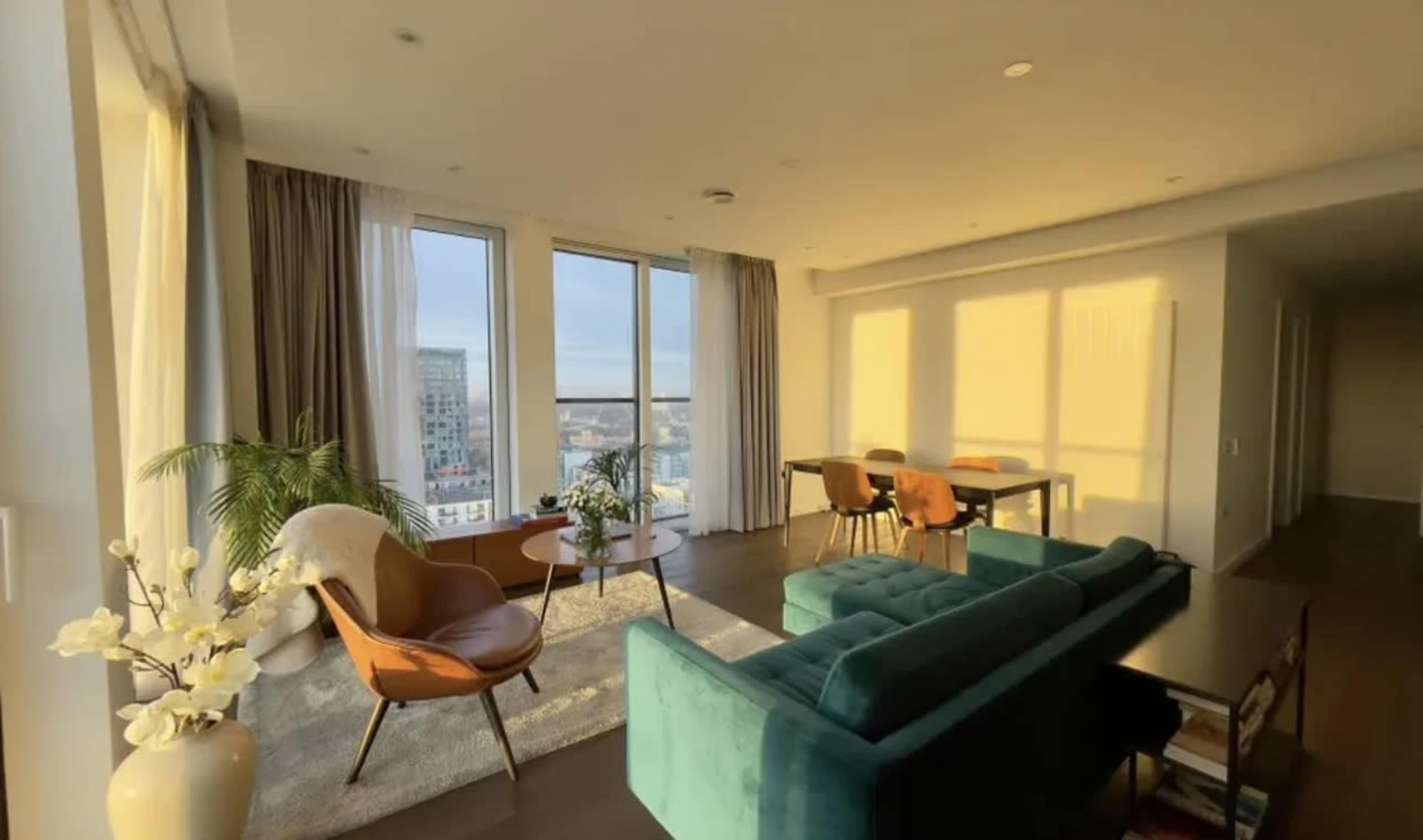 A modern living room features a teal sofa, orange chairs, a decorative plant, and large windows allowing natural light to illuminate the space.