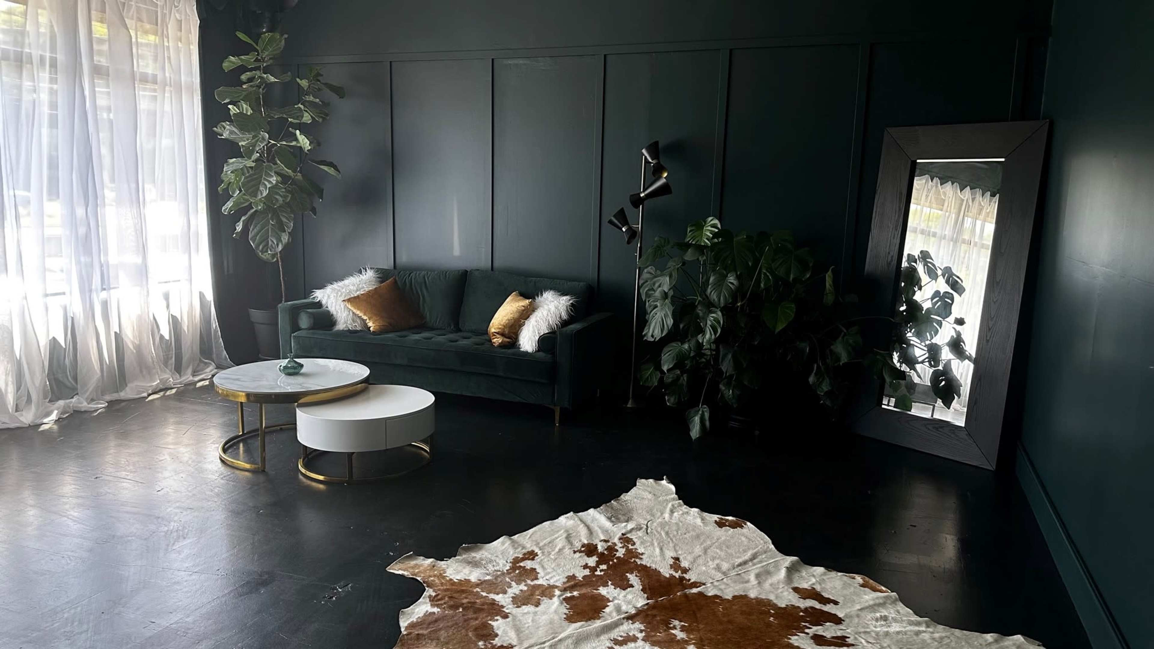 The room features a dark green wall with a green sofa, a round coffee table, a large mirror, a cowhide rug, and various plants.