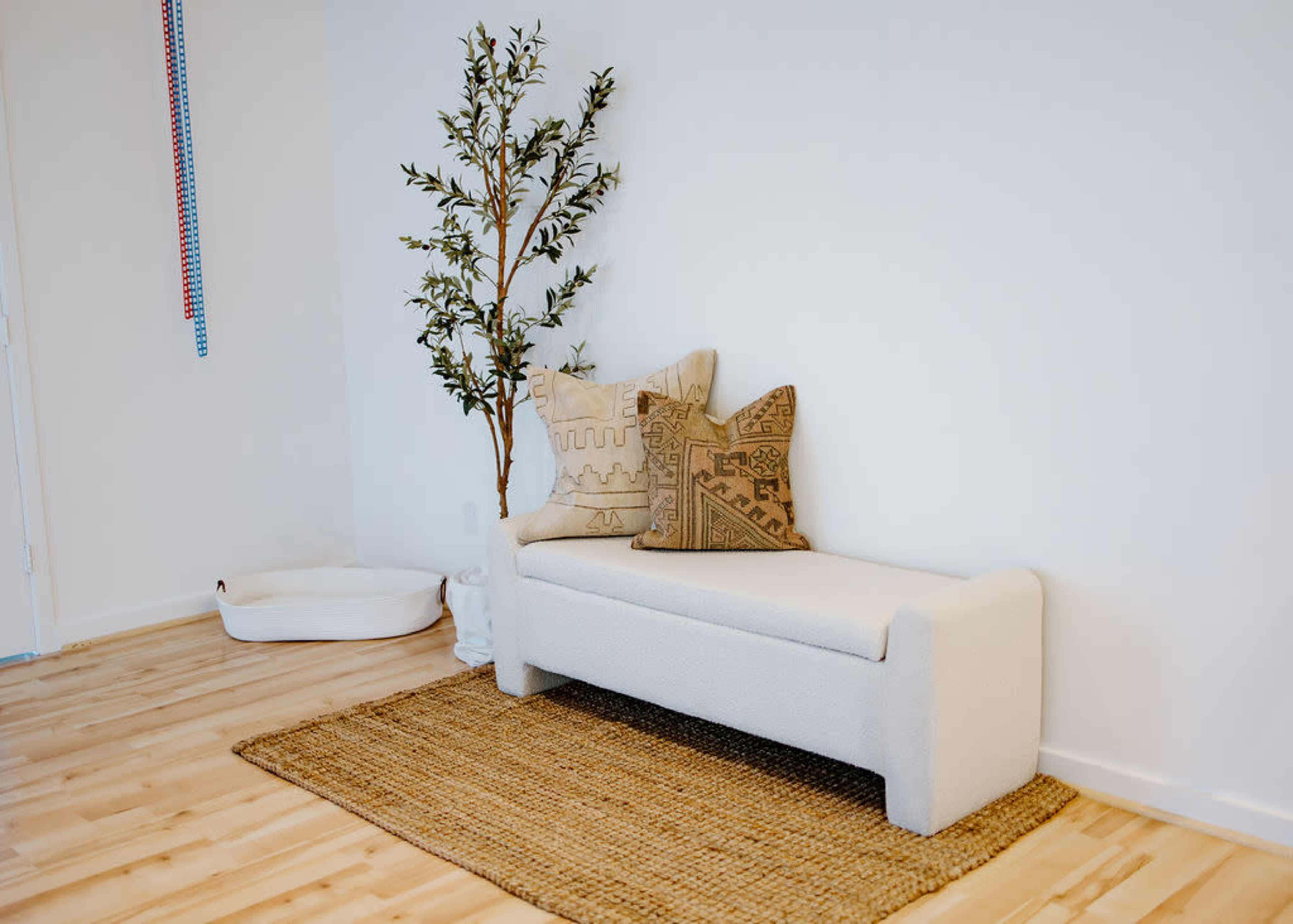 A minimalist living space featuring a light-colored sofa with decorative pillows, a small potted tree, and a woven rug.