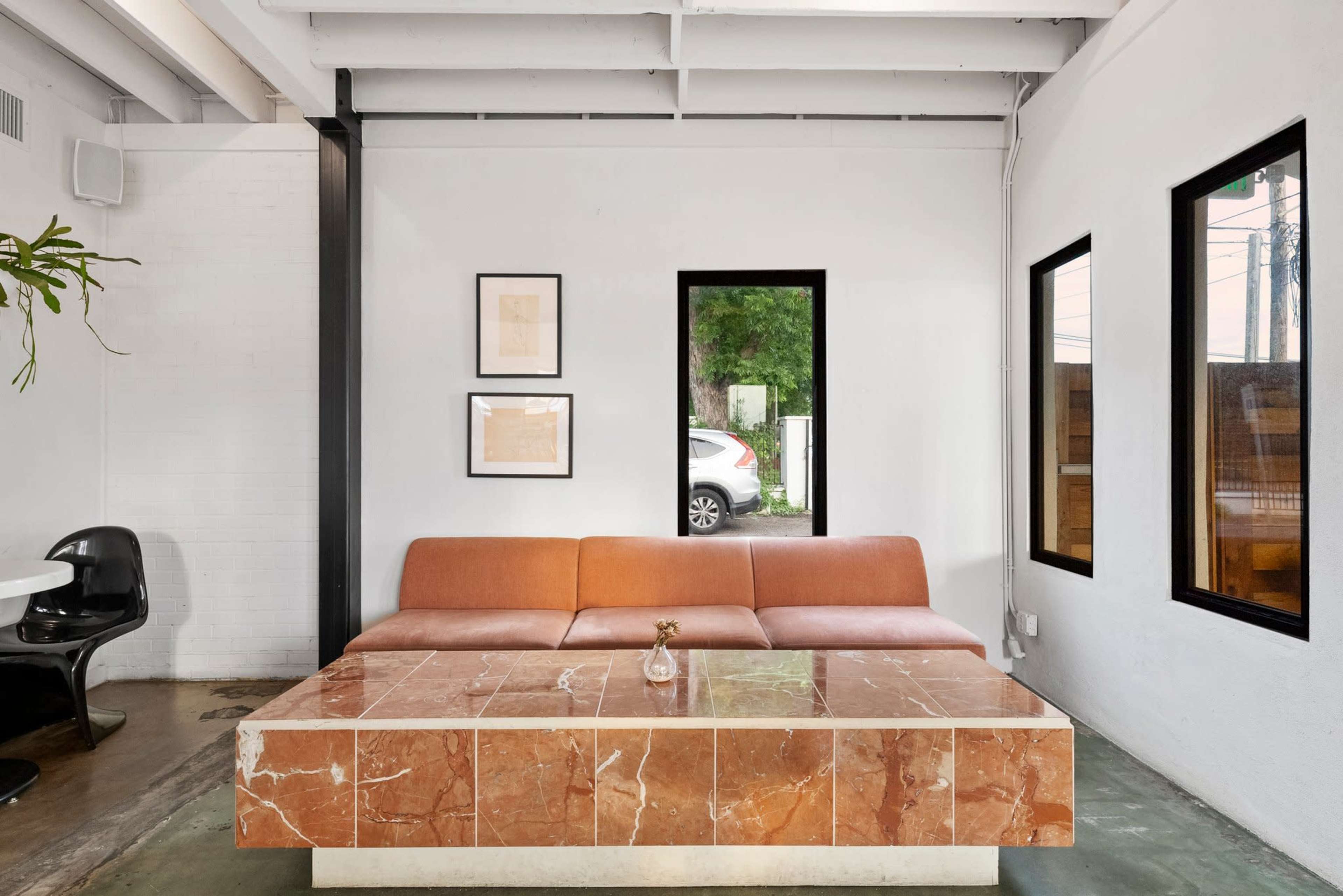 The image shows a modern interior space featuring a large brown sofa and a rectangular marble coffee table, with windows allowing natural light to enter.