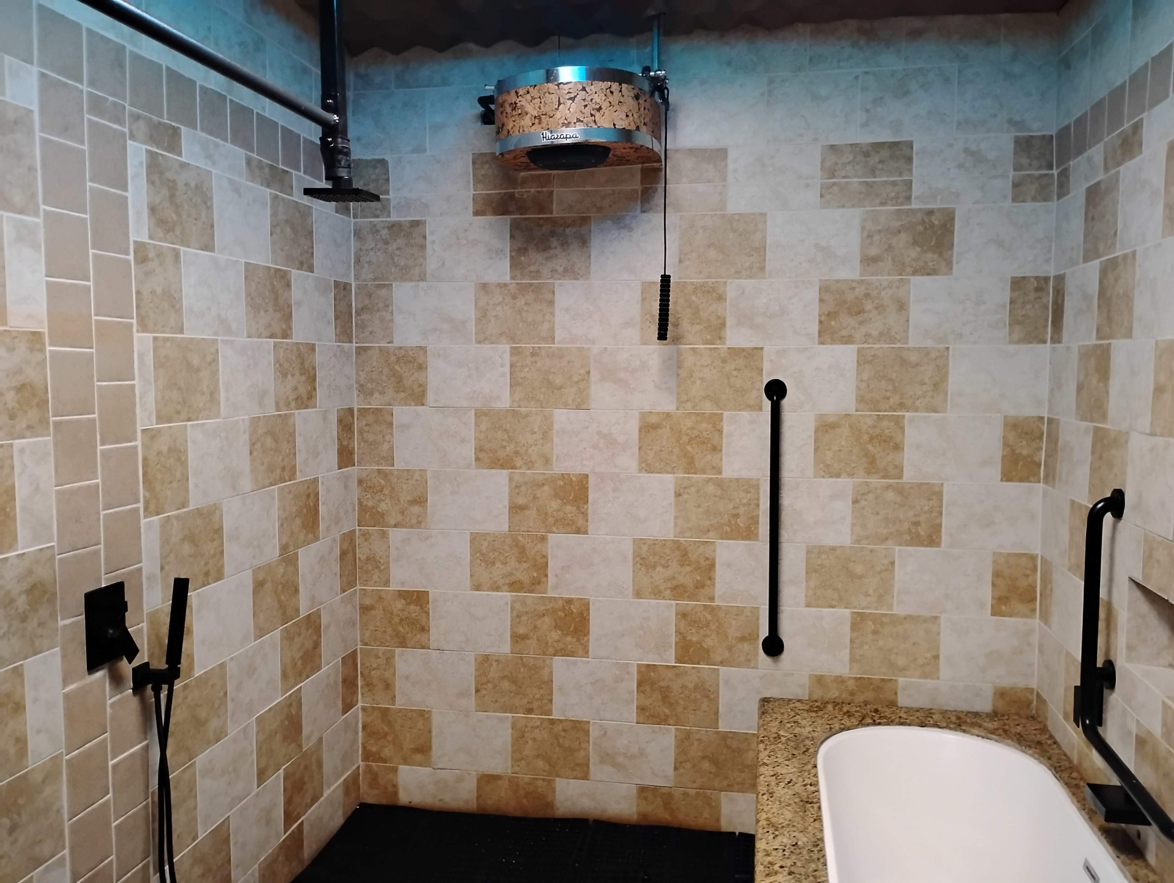 The image shows a modern bathroom with tiled walls, a round overhead shower, a bathtub, and black grab bars.