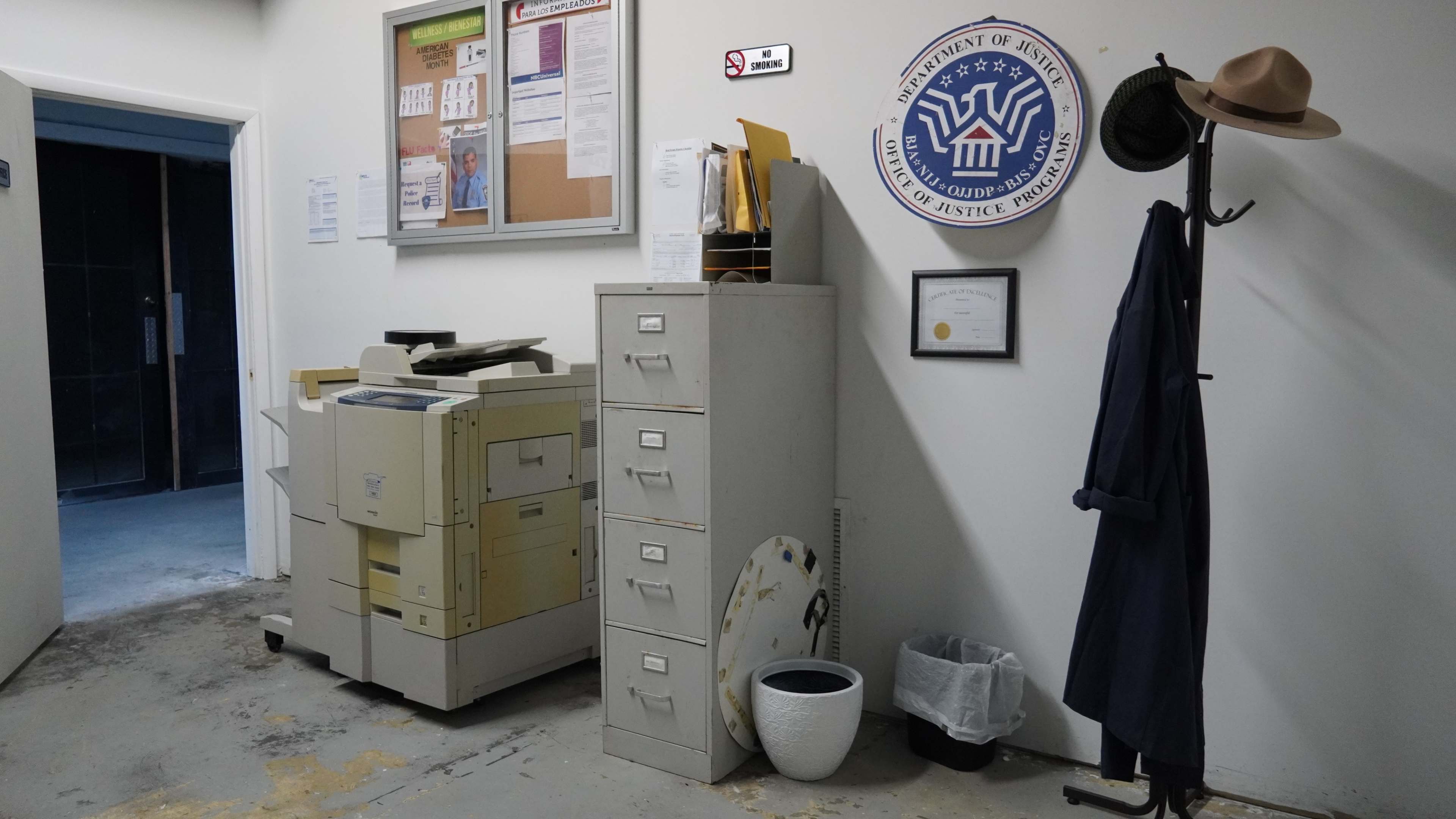 The image shows an office corridor with a copier, a filing cabinet, and a coat rack holding a hat and a coat.