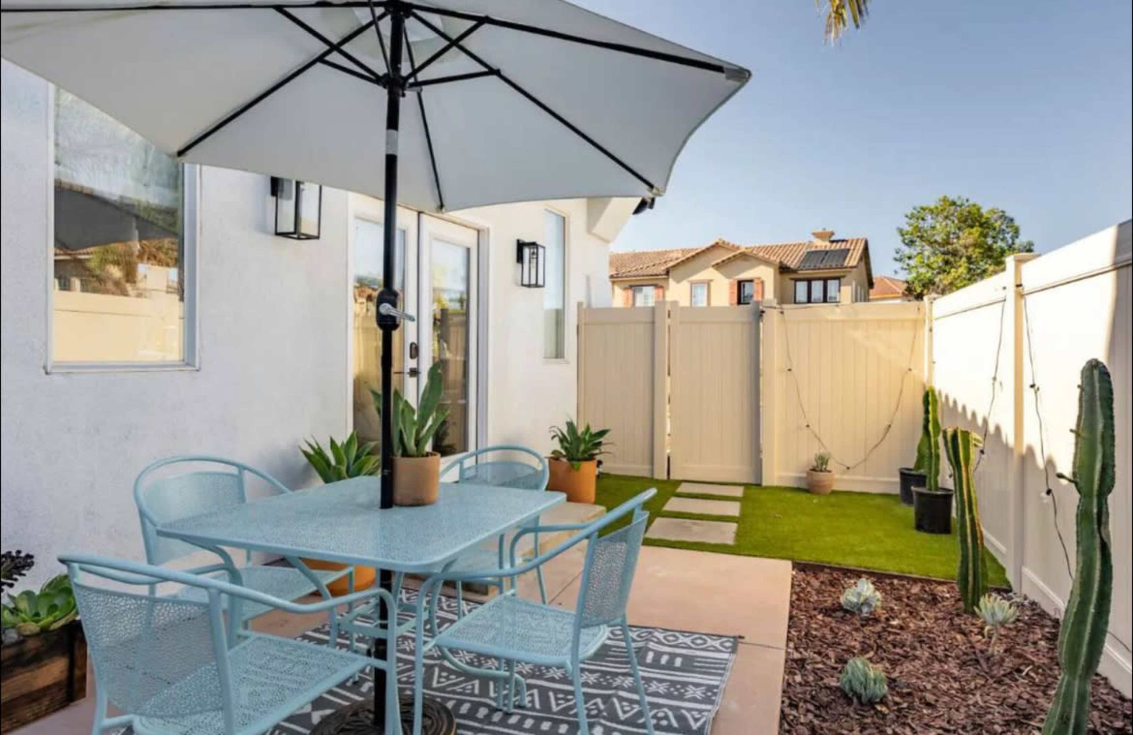 A patio features a table and chairs under an umbrella, surrounded by potted plants and a grass area enclosed by a fence.