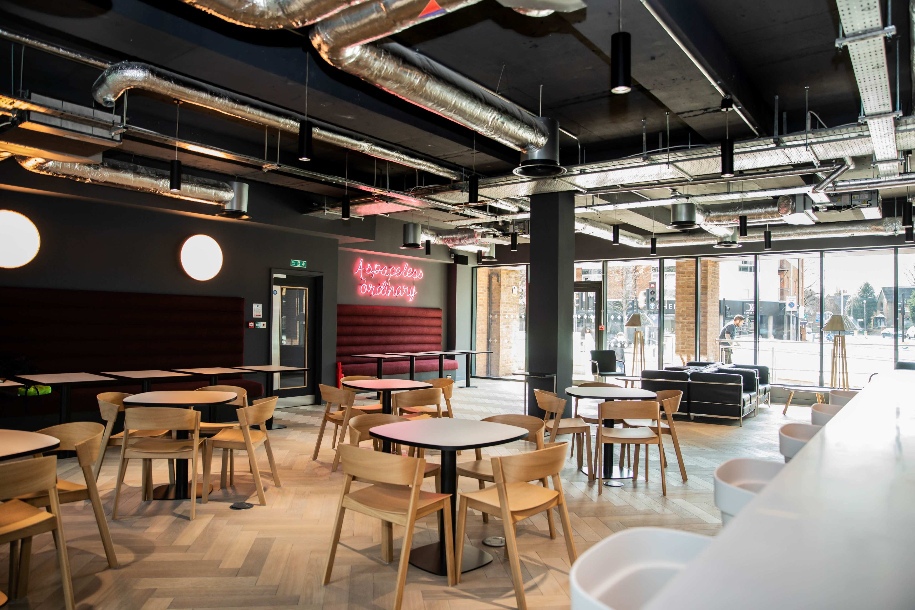 A modern cafe interior features wooden tables and chairs, large windows, and a neon sign on the wall.
