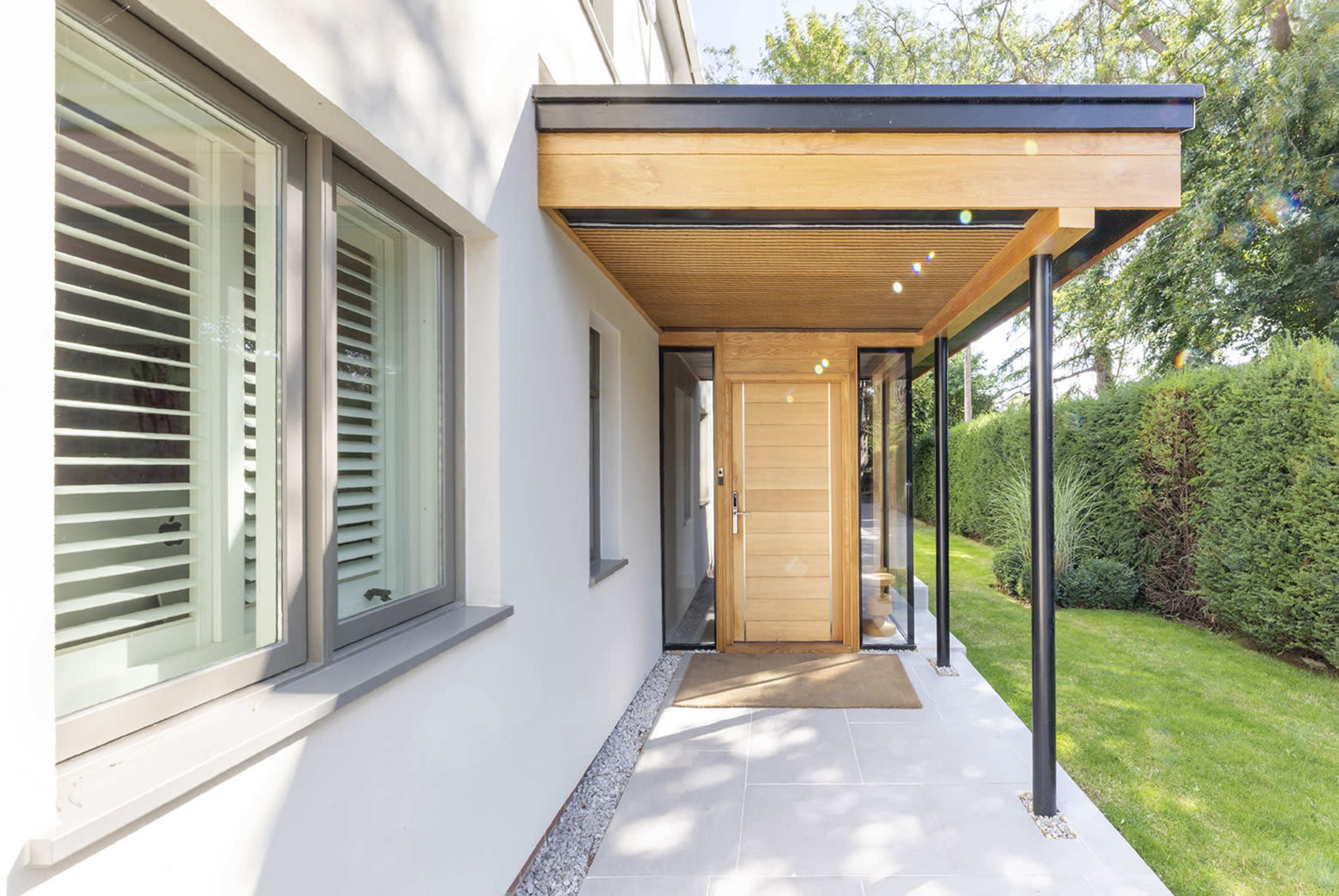 The image shows a modern entrance with a wooden door, flanked by large windows and a covered porch, leading to a pathway surrounded by greenery.