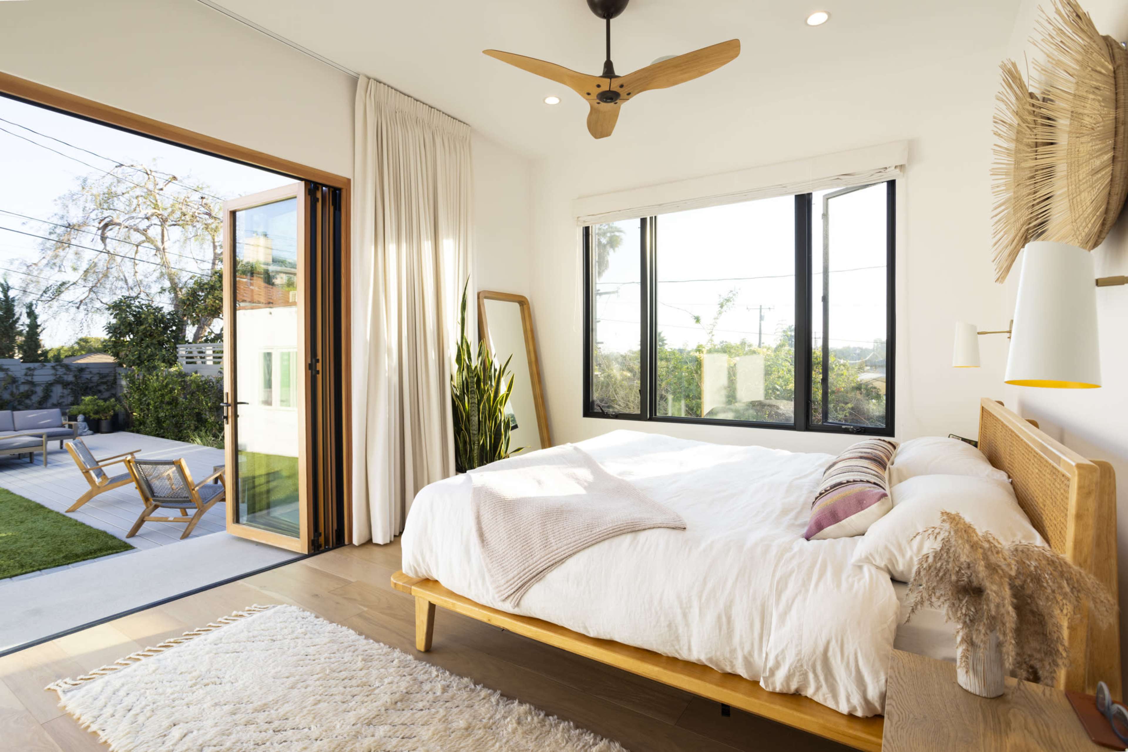 A bright bedroom features a large bed with a light bedding set, a ceiling fan, and an open door leading to a patio.