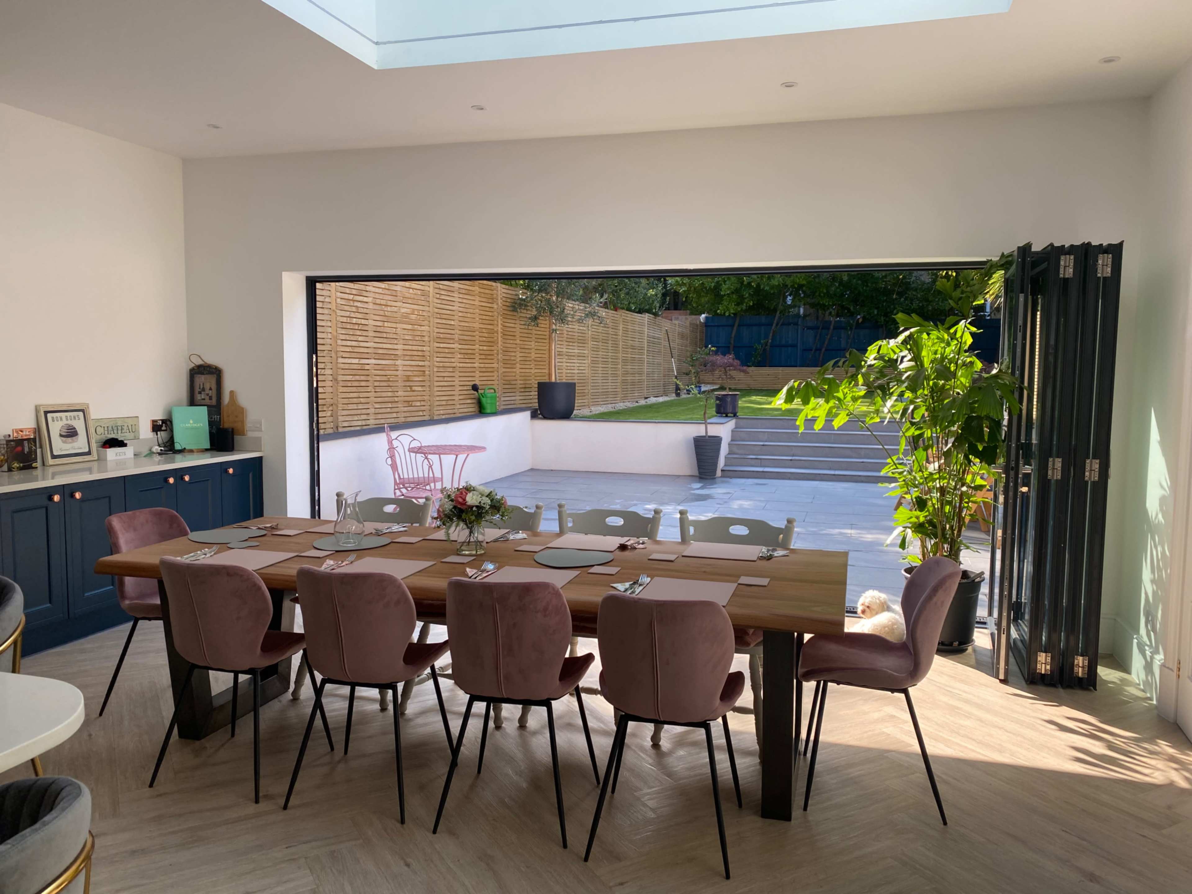 A spacious dining area features a large wooden table surrounded by pink upholstered chairs, with a view of an outdoor garden through open glass doors.