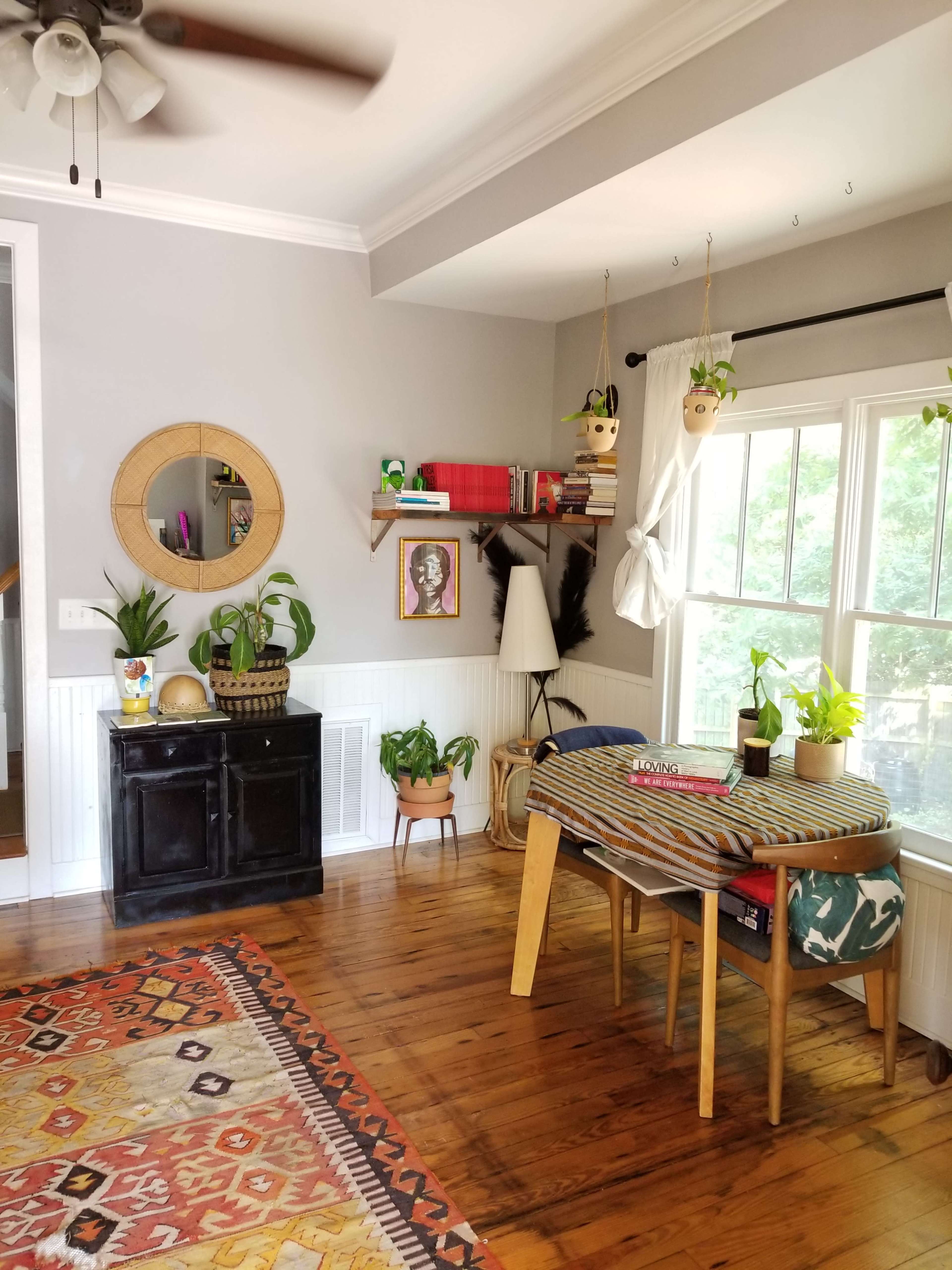 The scene features a cozy room with wooden flooring, a black cabinet, a window with plants, and a small table stacked with colorful items.