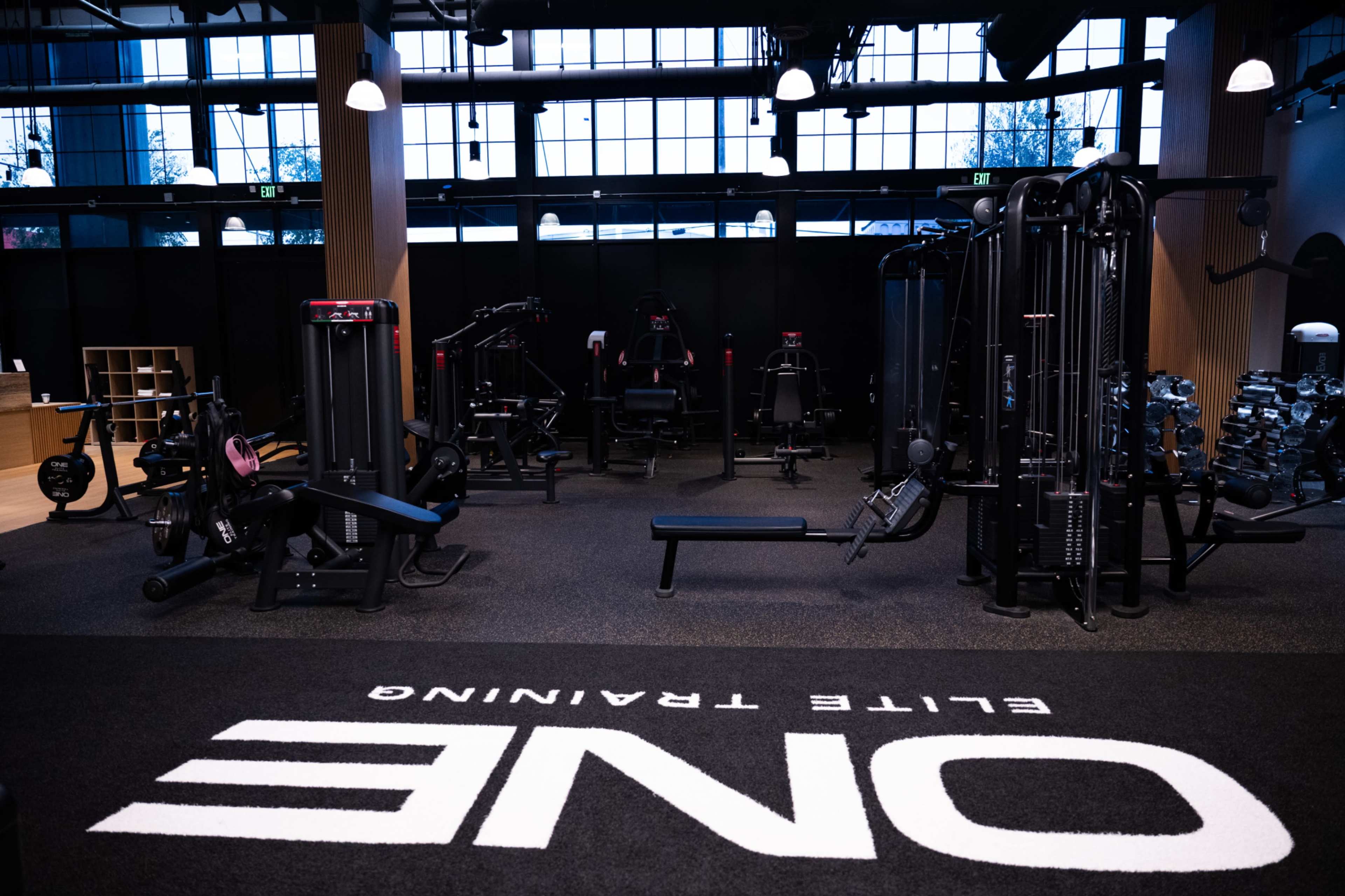 The image shows a modern gym interior featuring various strength training equipment and exercise machines arranged on a carpeted floor with the word "ONE" prominently displayed.