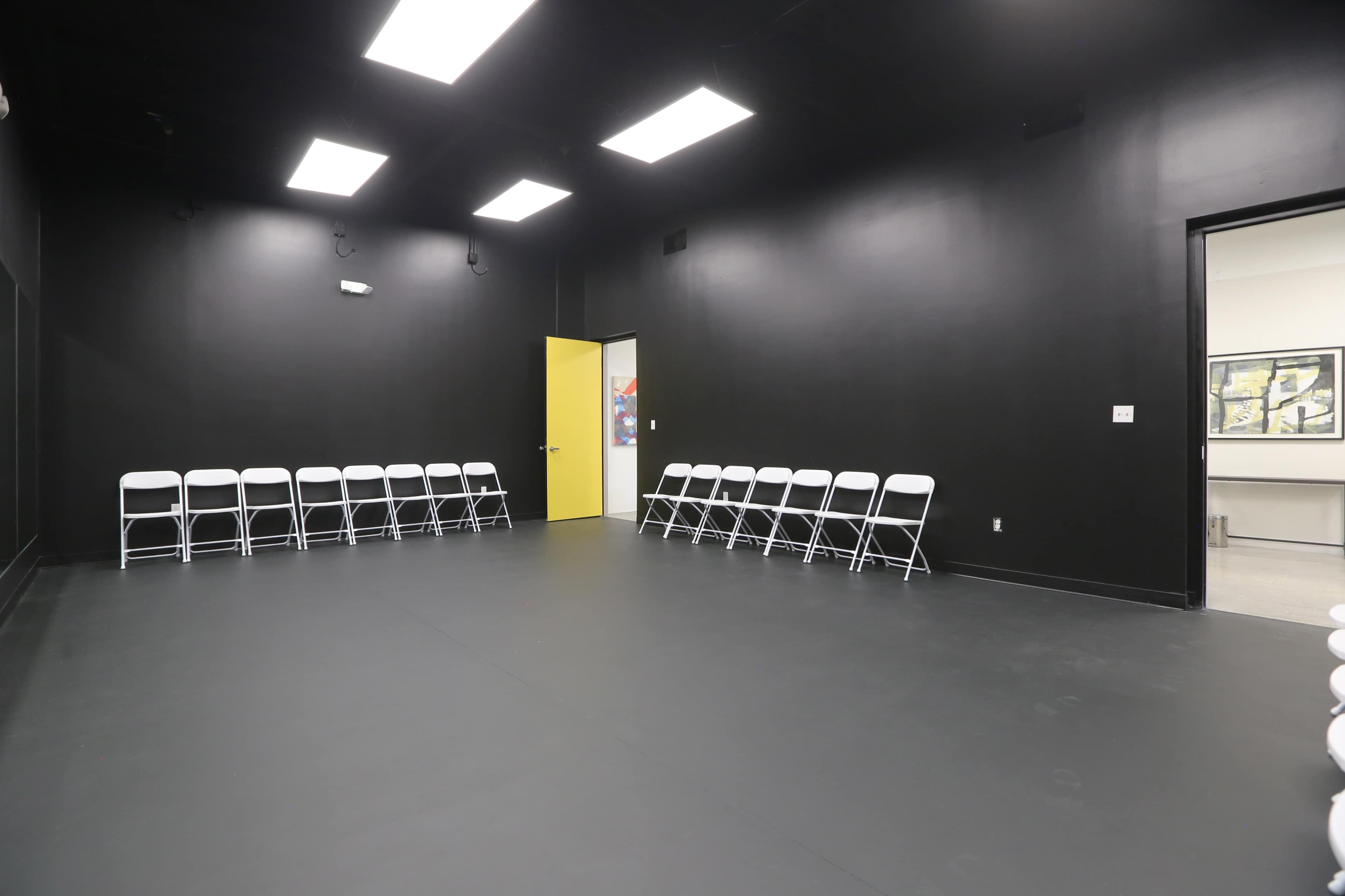 A stark black-walled room contains a single line of white folding chairs facing a yellow door.