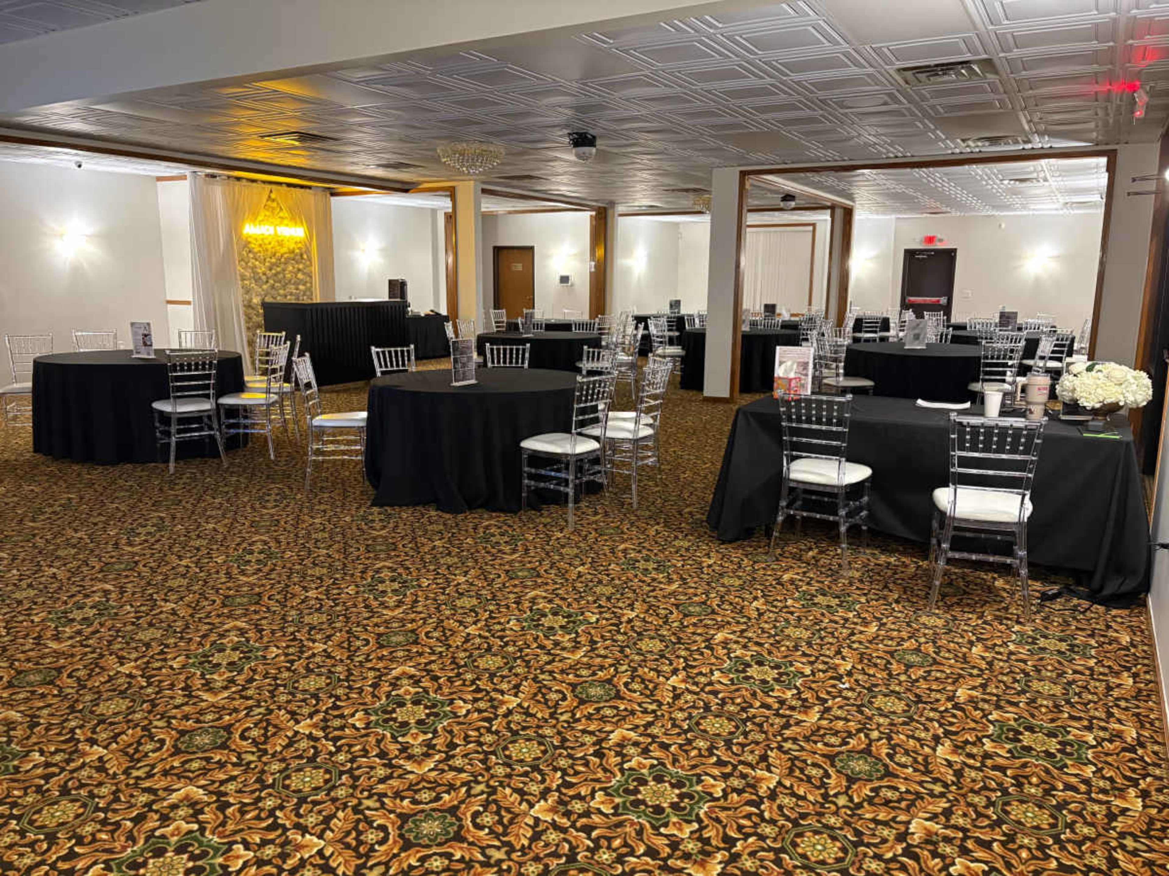 The image shows a banquet hall set up with several round tables covered in black tablecloths and clear acrylic chairs, featuring a patterned carpet and illuminated signage in the background.