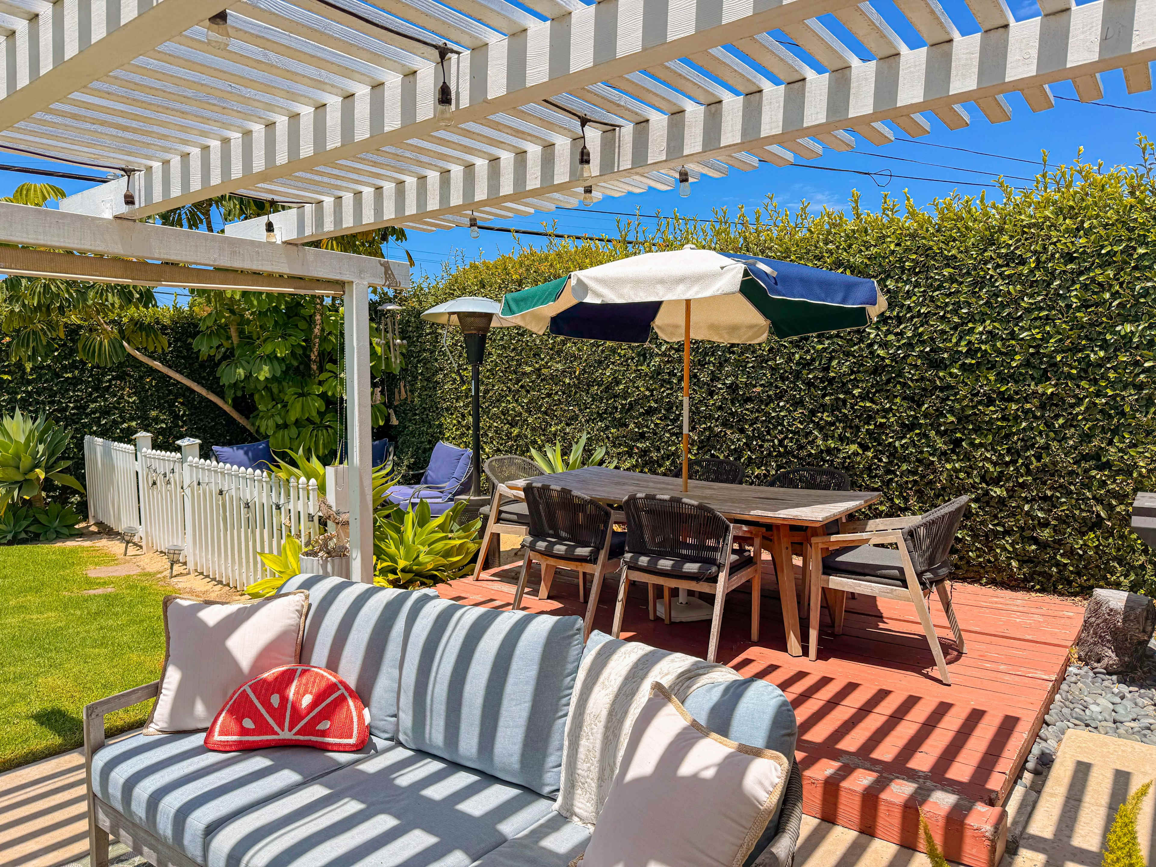 The image shows a patio area with a dining table under an umbrella, surrounded by greenery and furnished with a couch and decorative pillow.