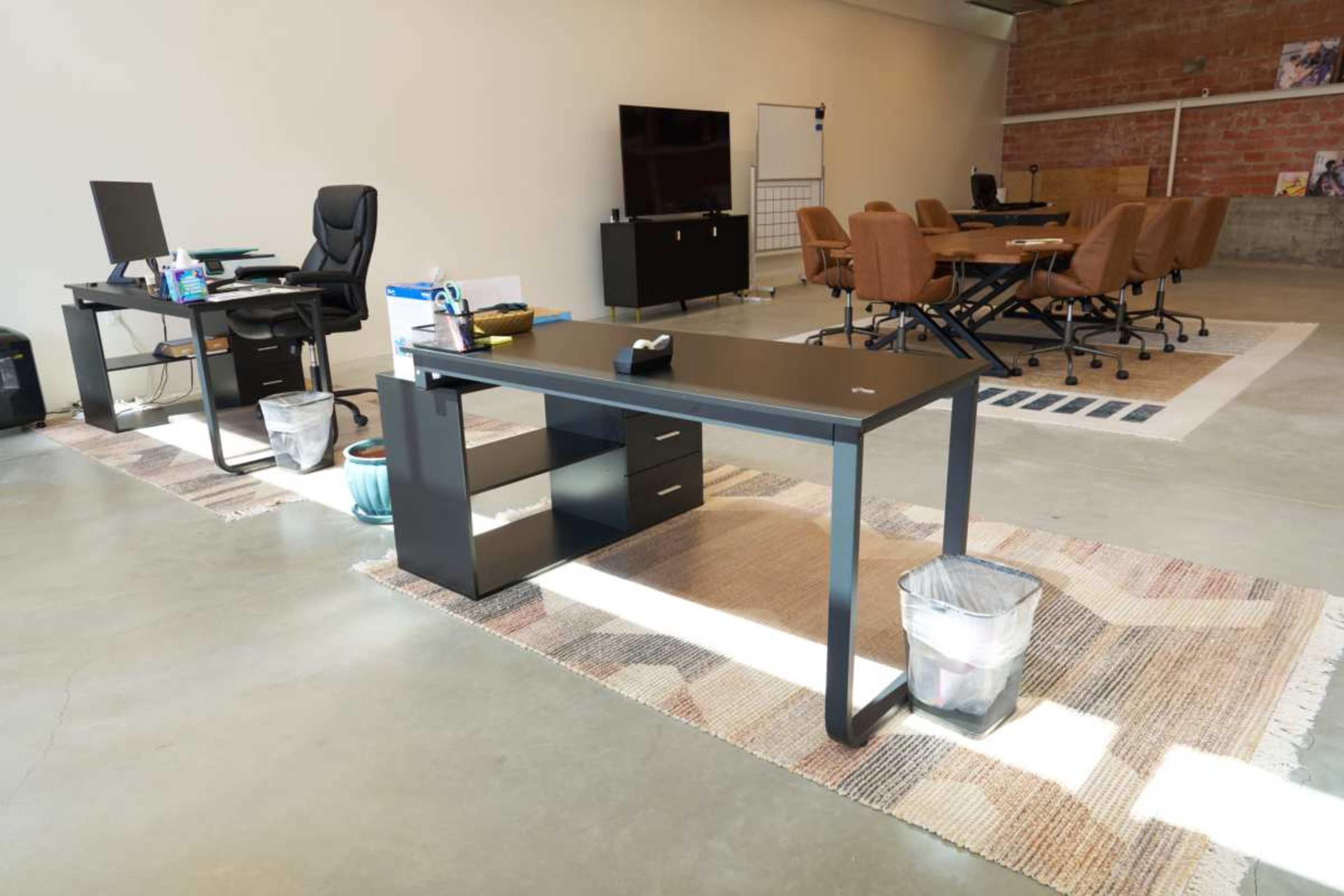 The image shows a modern office space featuring two desks, a black conference table with chairs, and a carpeted area on a concrete floor.