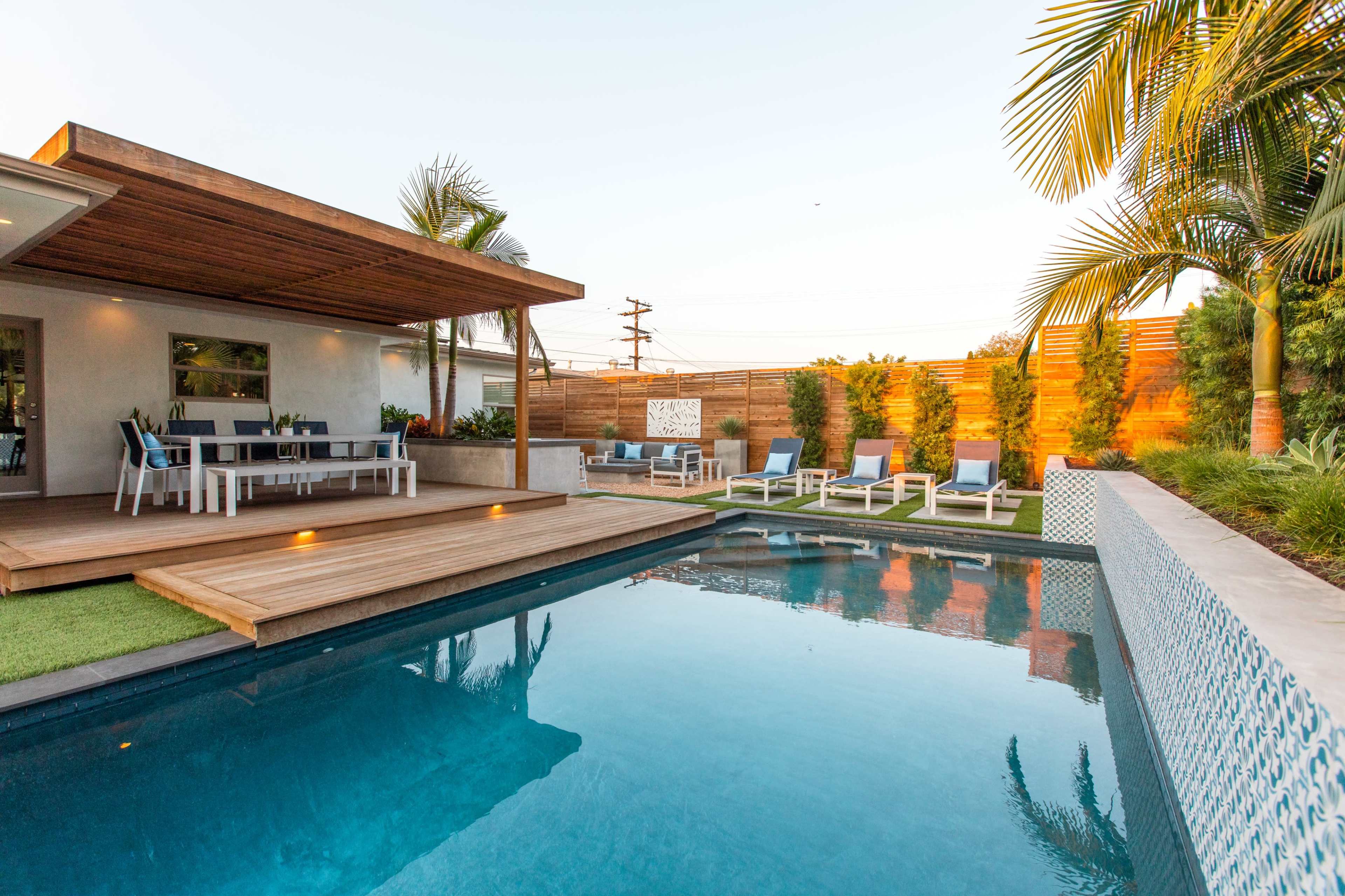 A modern backyard features a pool surrounded by lounge chairs and a wooden deck with a patio area for dining.