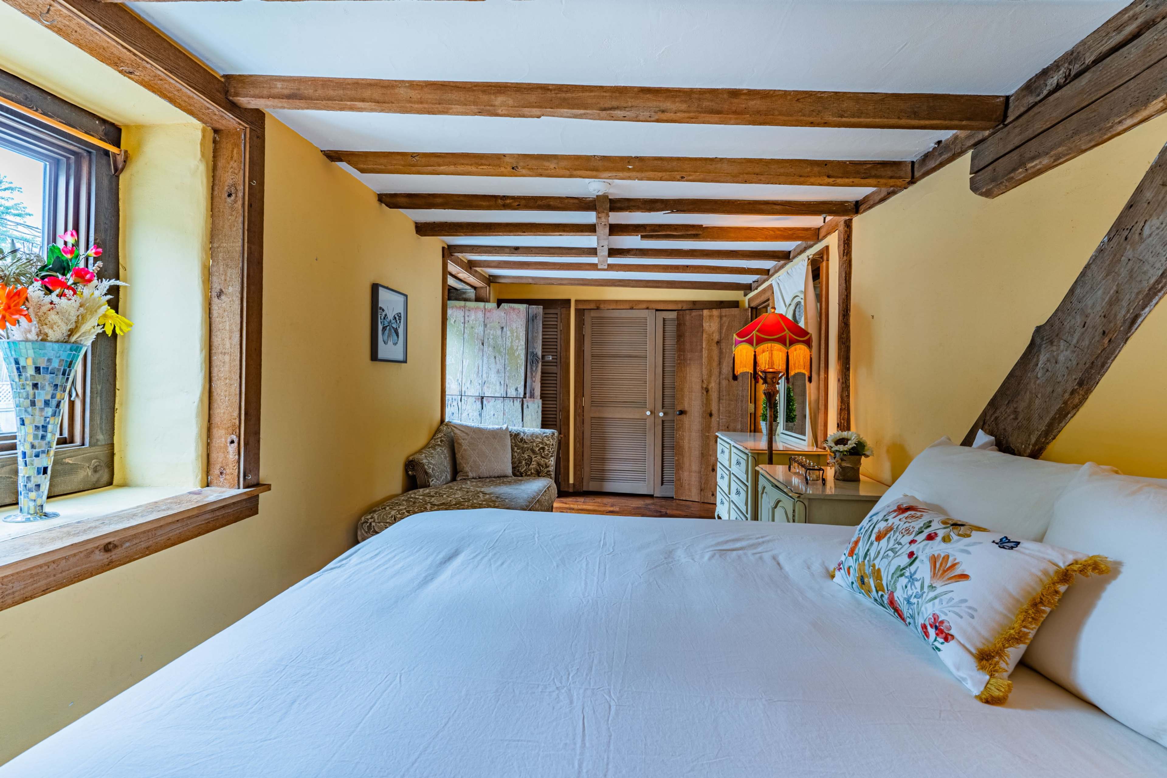 A cozy bedroom features a large bed, exposed wooden beams, and a sitting area by the window with a decorative vase of flowers.