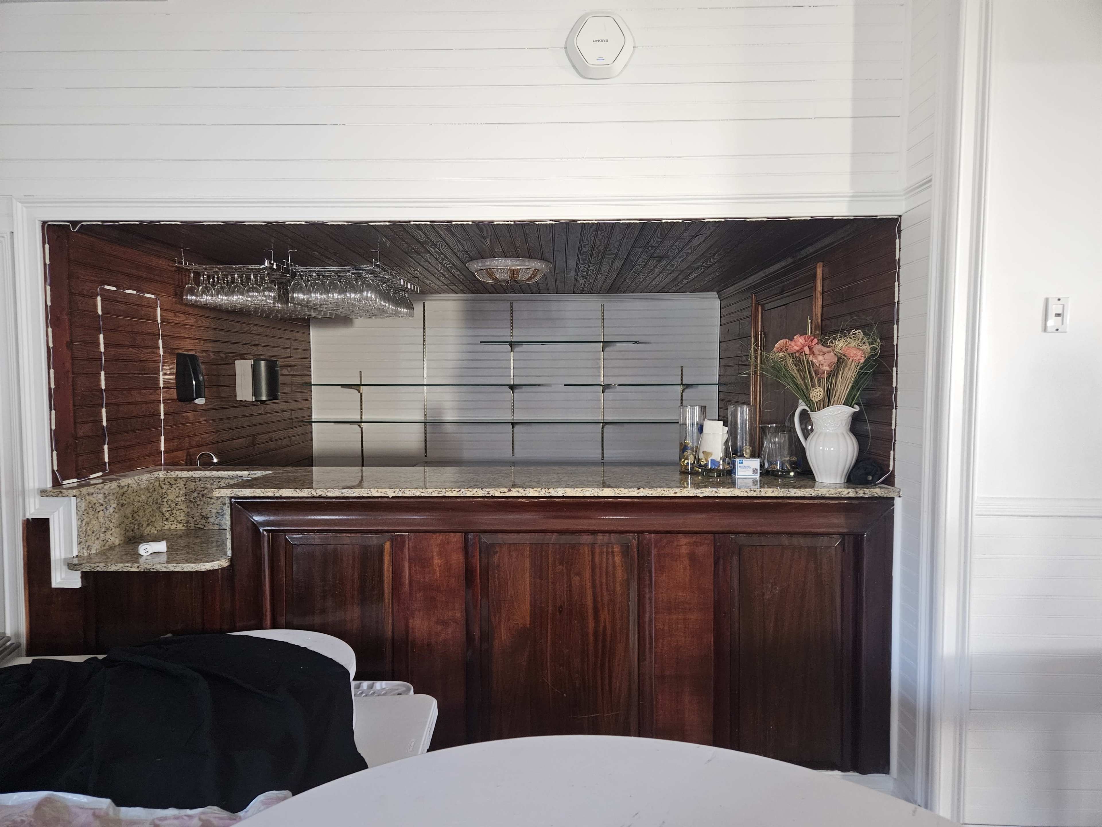 The image shows a wooden bar area with a granite countertop, flanked by a set of shelves and an arrangement of glassware and a vase with flowers in the background.