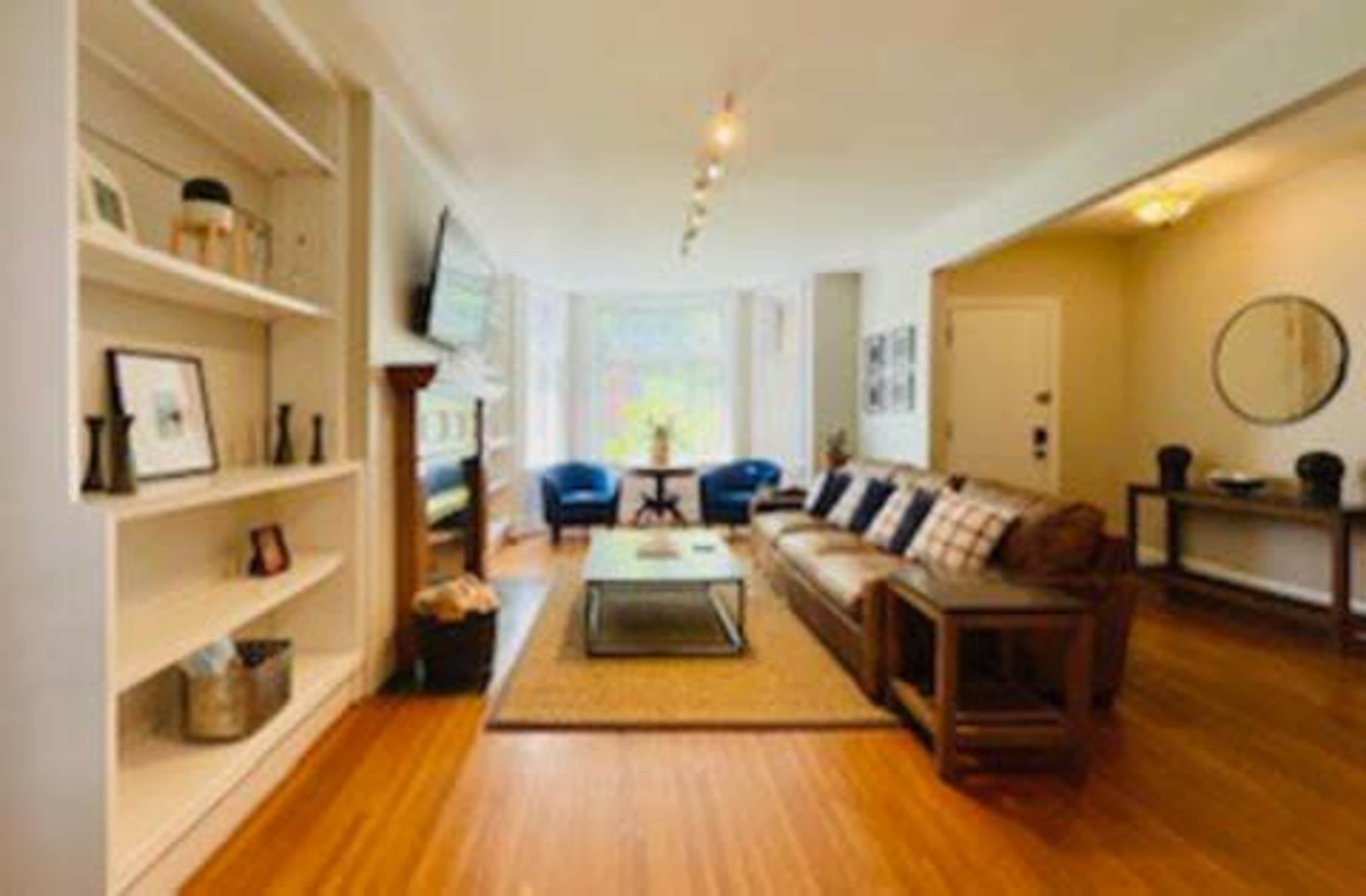 The image shows a modern living room with a brown sofa, blue chairs, a coffee table, and bookshelves against the walls.