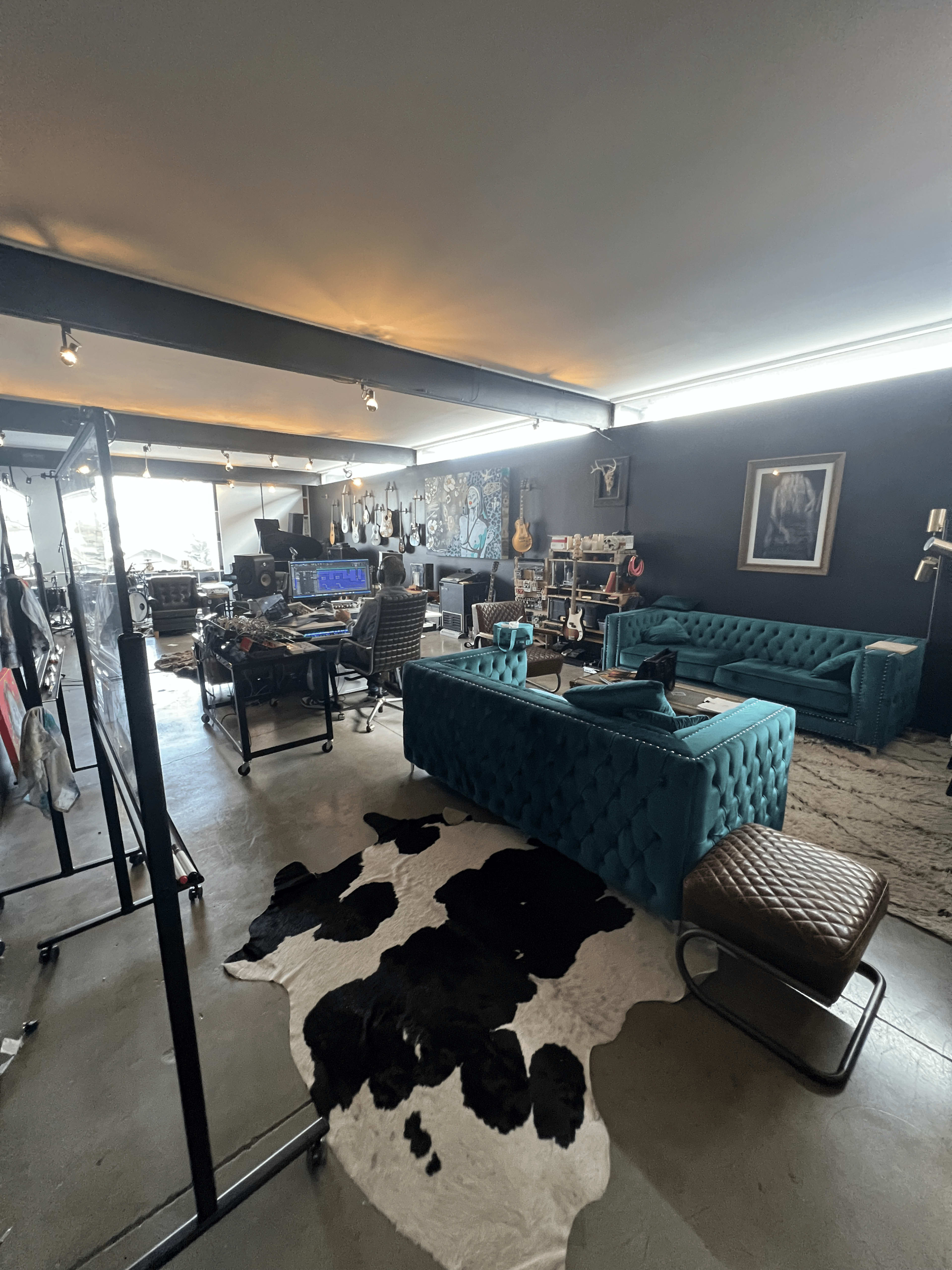 The image shows a spacious studio with turquoise tufted sofas, a cowhide rug on the floor, musical instruments on display, and various workstations set up for creative activities.