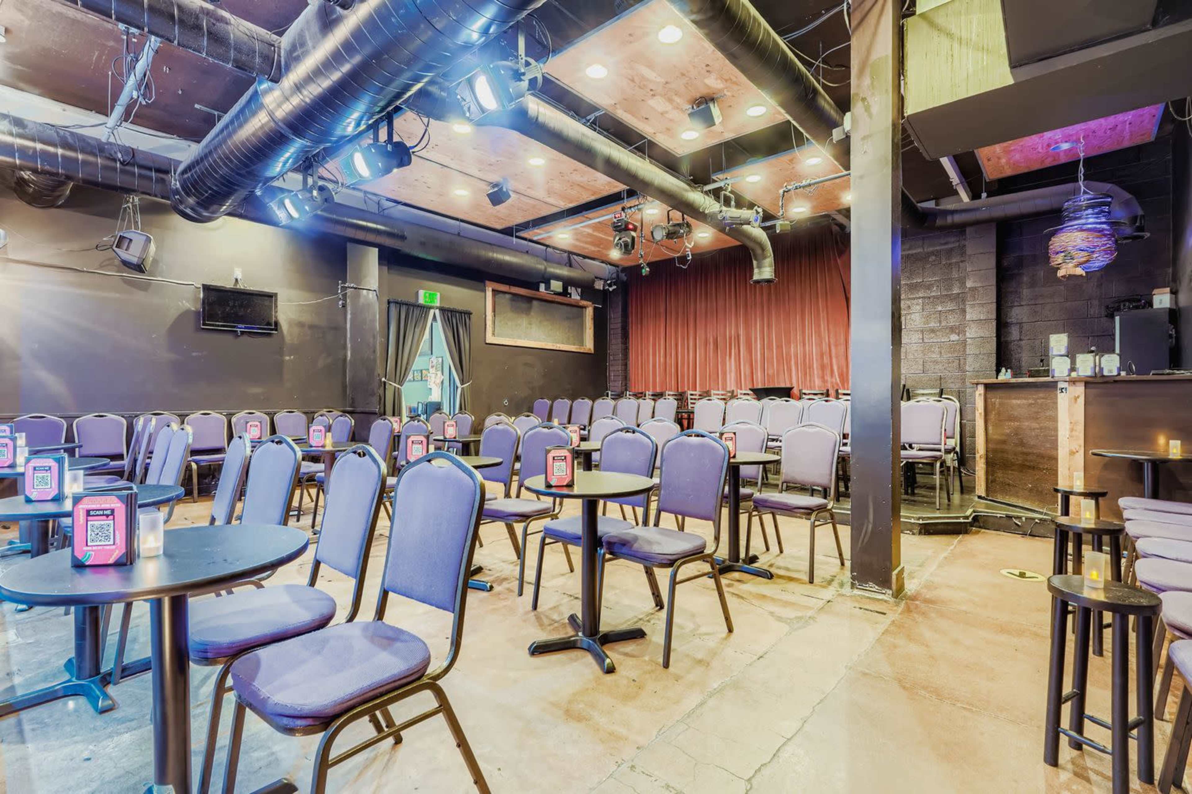The image shows an empty performance venue with rows of chairs, small tables, and stage lighting.