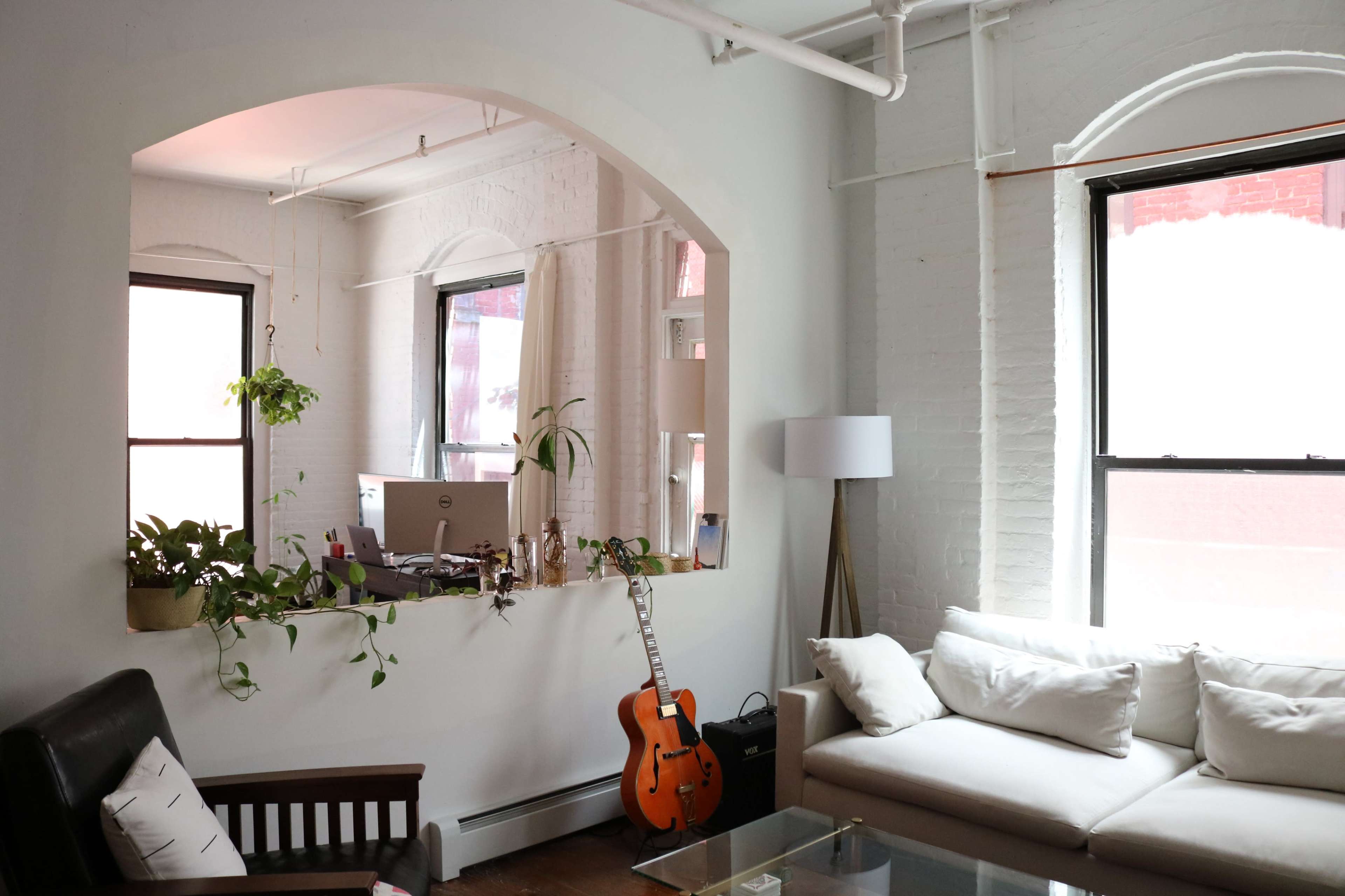 A well-lit living space with an archway leading to an office area, featuring a light-colored sofa, a guitar resting against the wall, and indoor plants.
