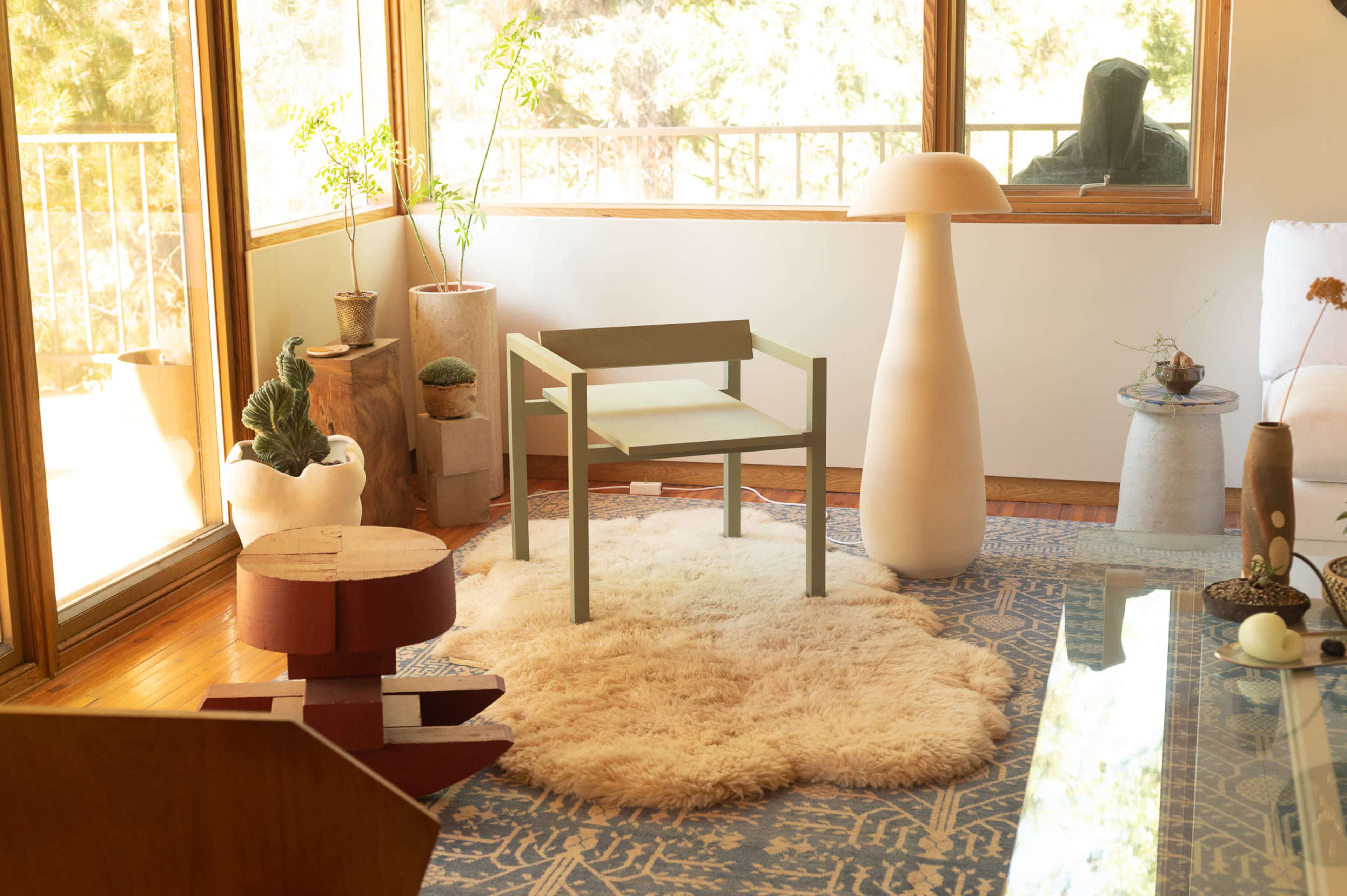 The scene features a modern interior with a minimalist chair, a sculptural lamp, and a decorative rug, surrounded by plants and natural light from large windows.