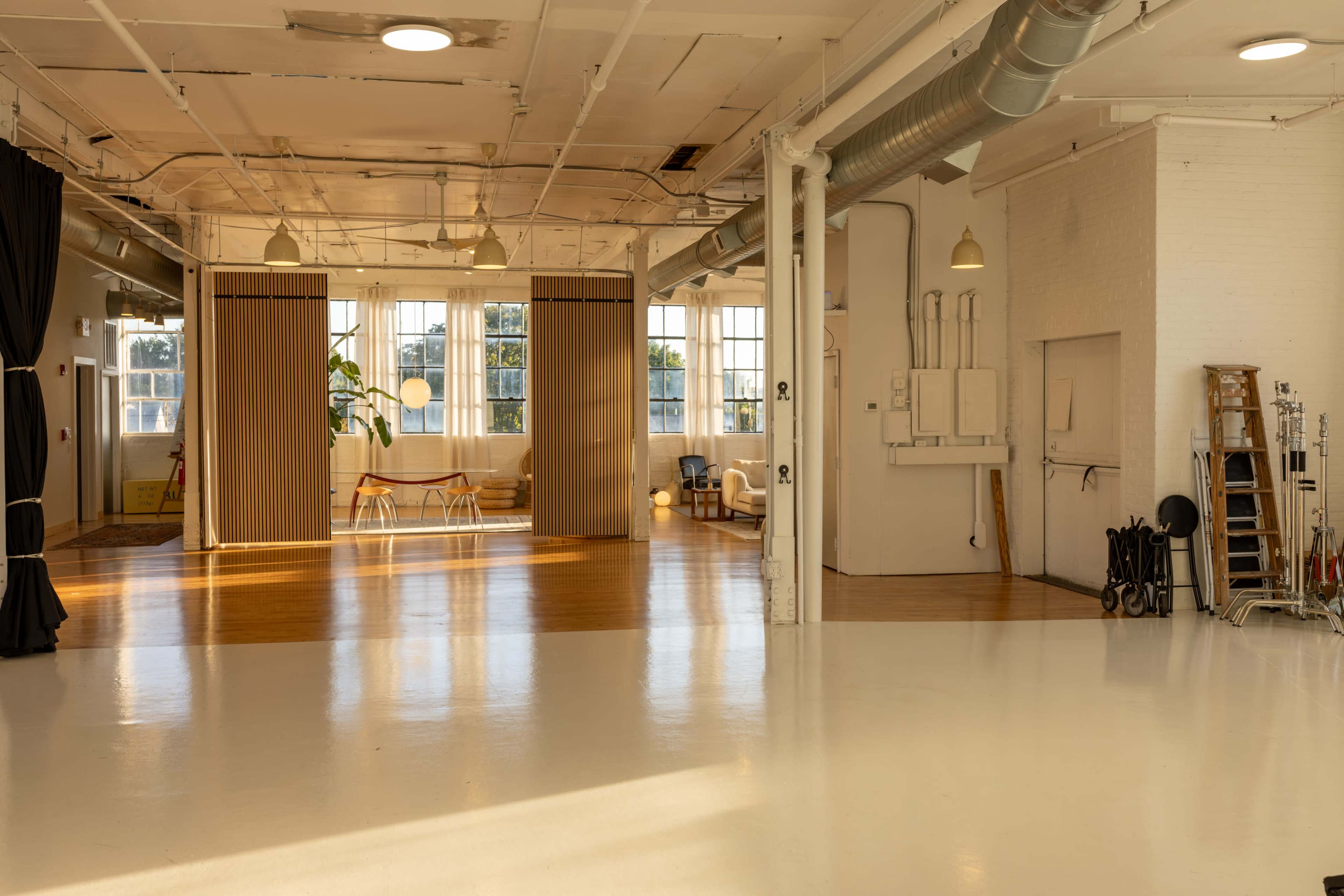 The image shows a spacious, well-lit studio with polished white floors, large windows, and furniture arranged in a minimalist style.