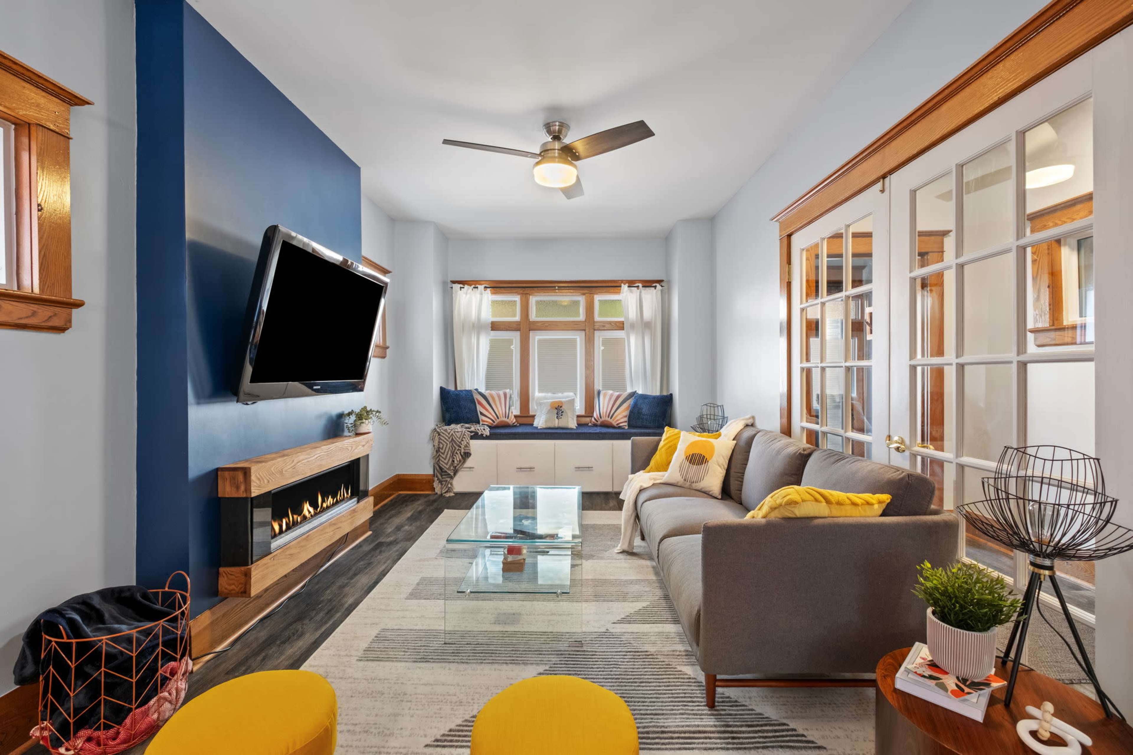 The image shows a modern living room with a gray sofa, a glass coffee table, and a mounted television on a blue feature wall, complemented by a cozy fireplace and bright window seating.