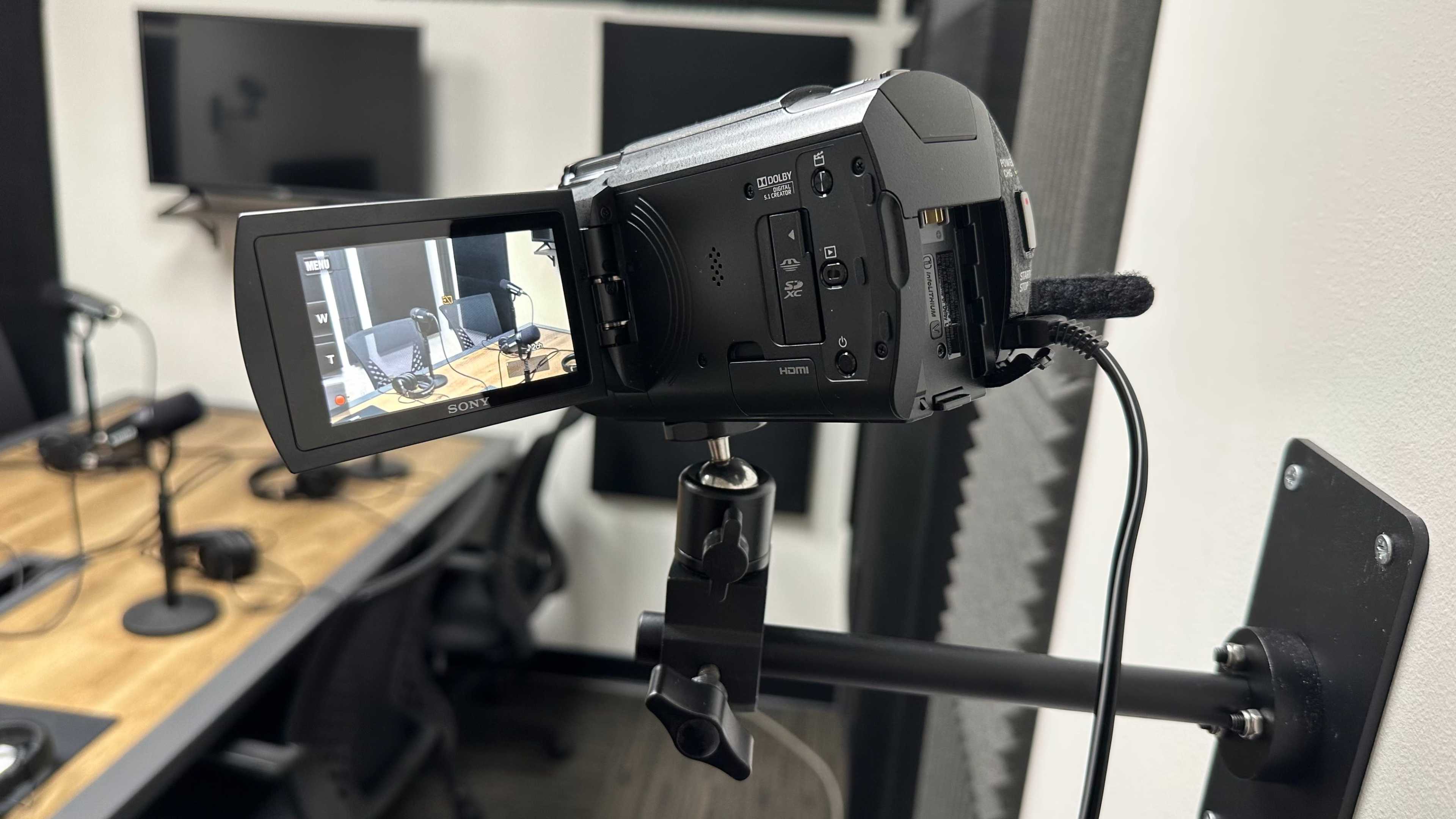 The image shows a Sony camcorder mounted on a tripod, facing a set of empty chairs and a table in a recording studio.