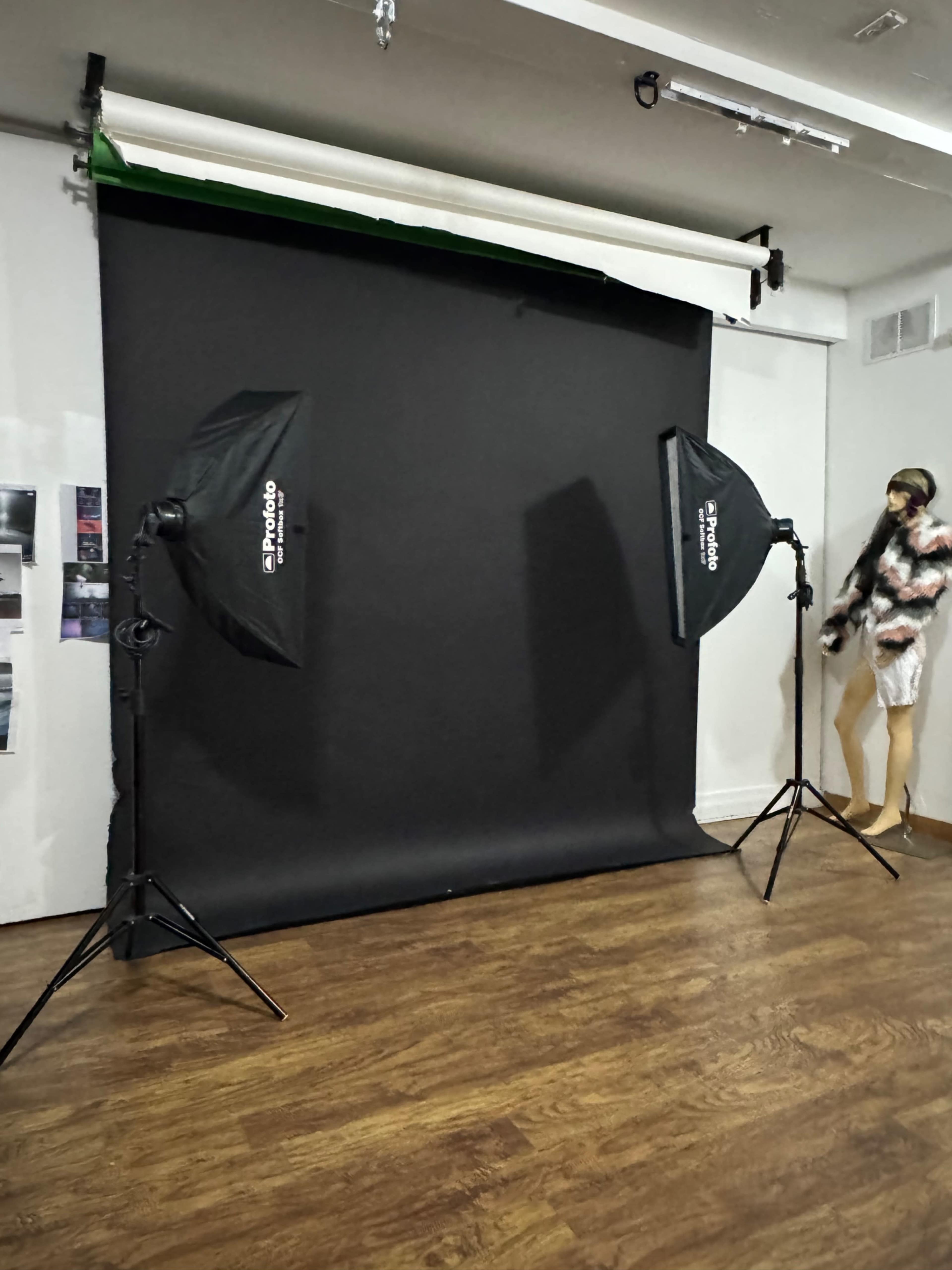 A photo studio setup features two softbox lights on stands flanking a black backdrop, with a mannequin dressed in a fur coat positioned nearby.
