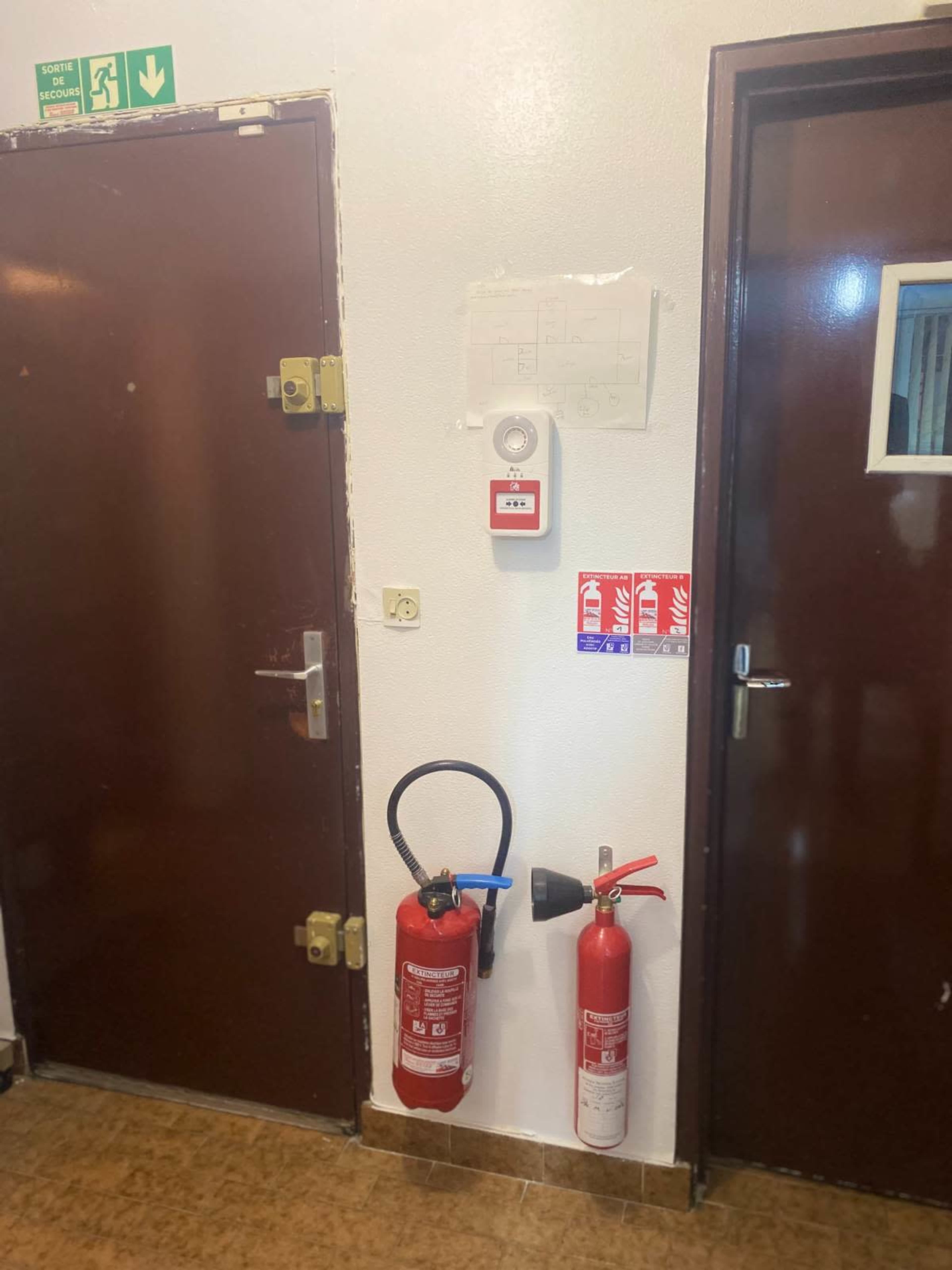 The image shows a wall with two fire extinguishers, a fire alarm, and doorways leading to two different rooms.