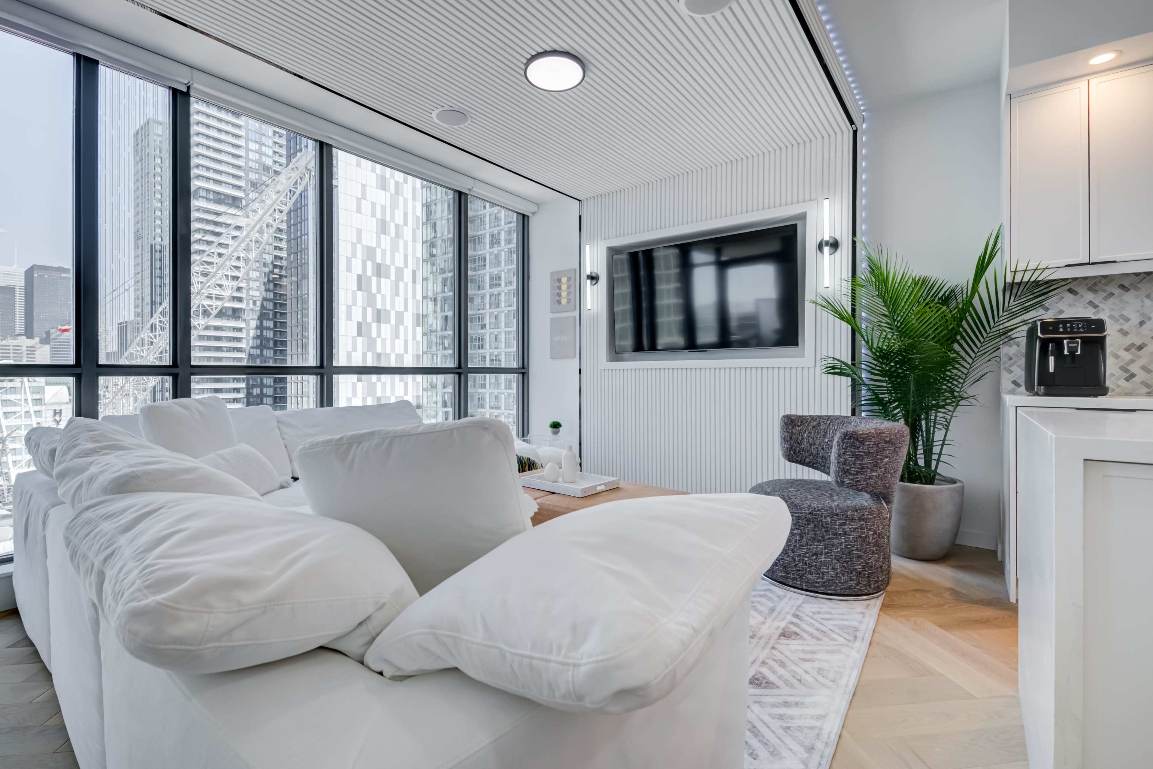 The image shows a modern living room with large windows, a sectional sofa, a small armchair, and a television mounted on the wall.