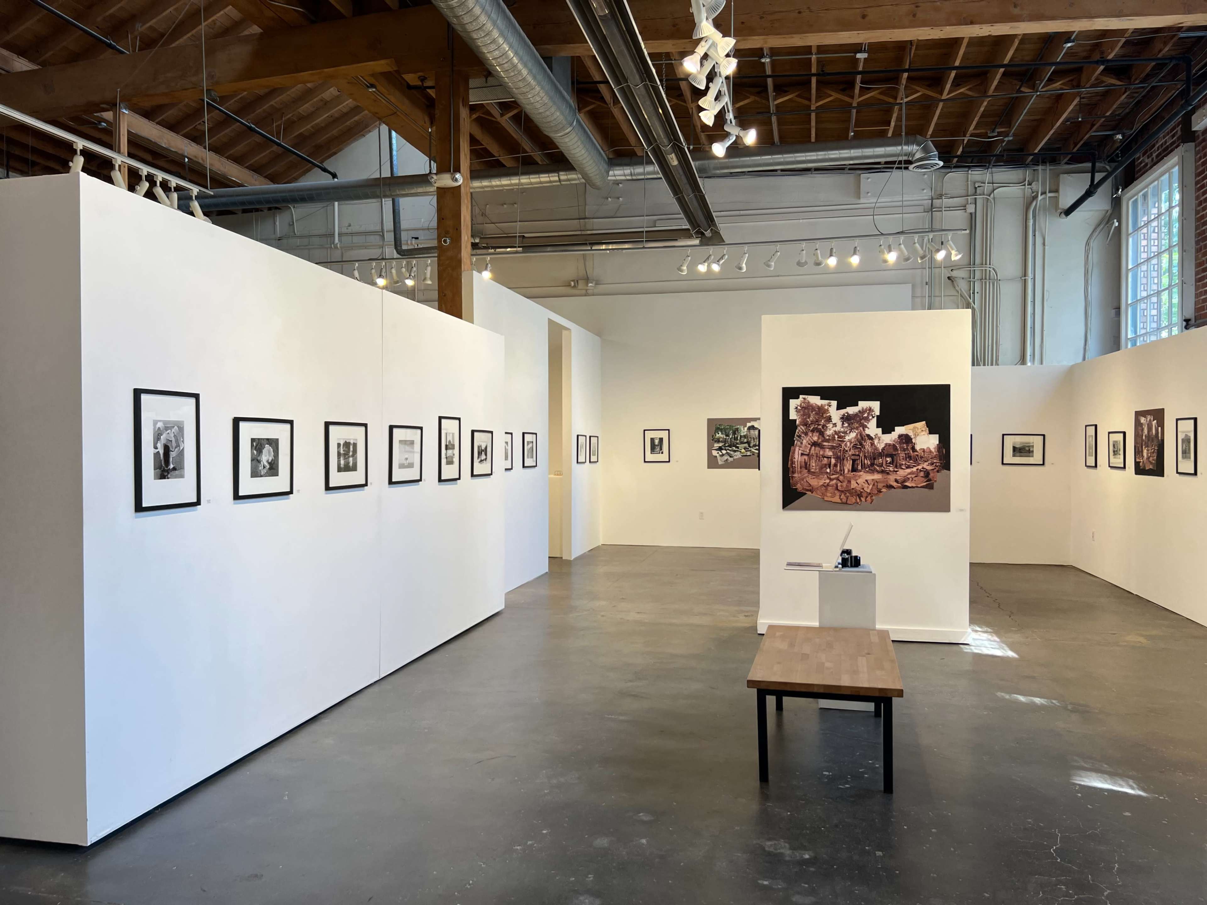 The image shows a spacious art gallery featuring white walls adorned with framed photographs, with a central wooden table.