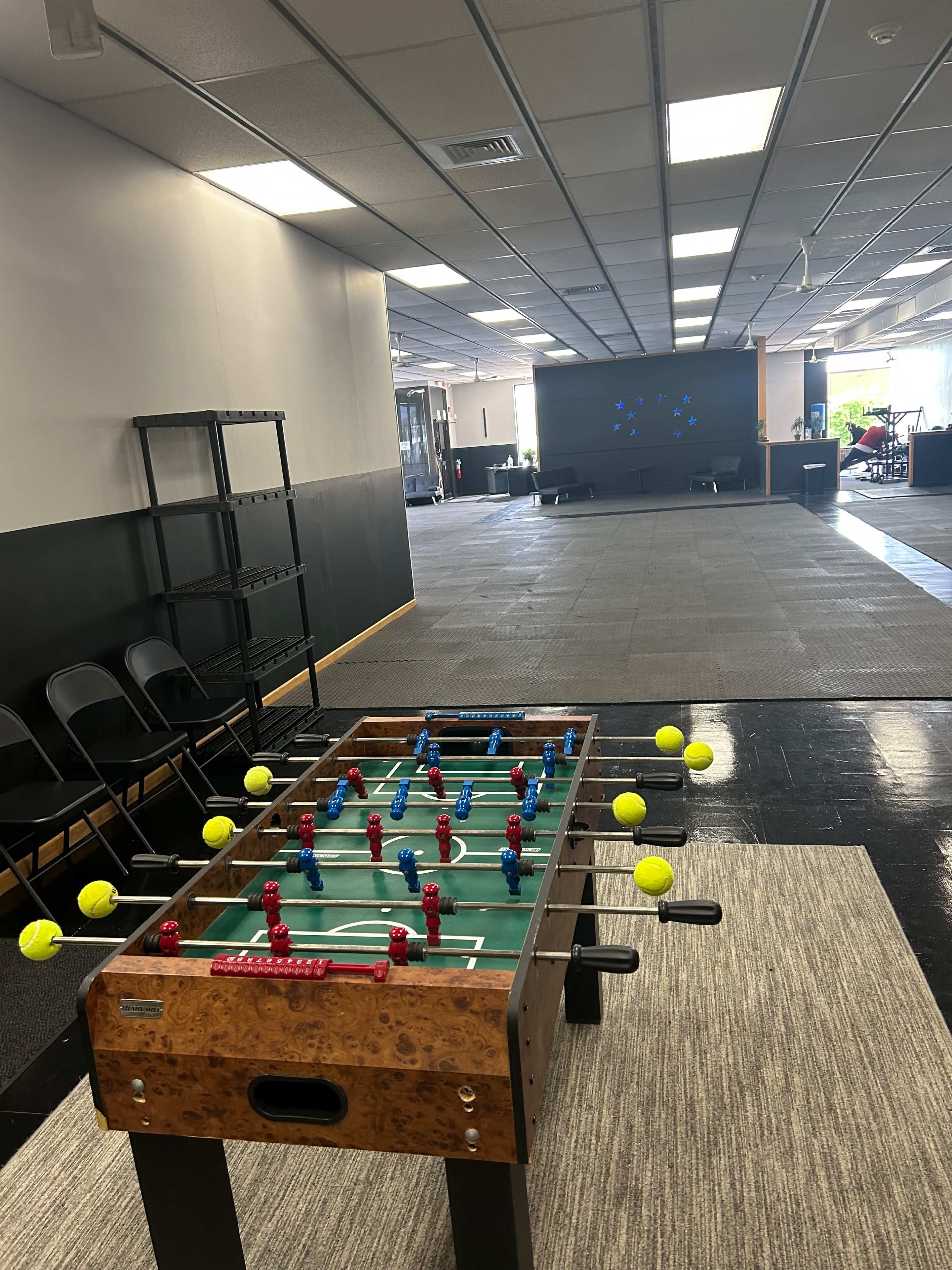 A foosball table with red and blue players is positioned in a spacious room, leading towards a large screen on the far wall.