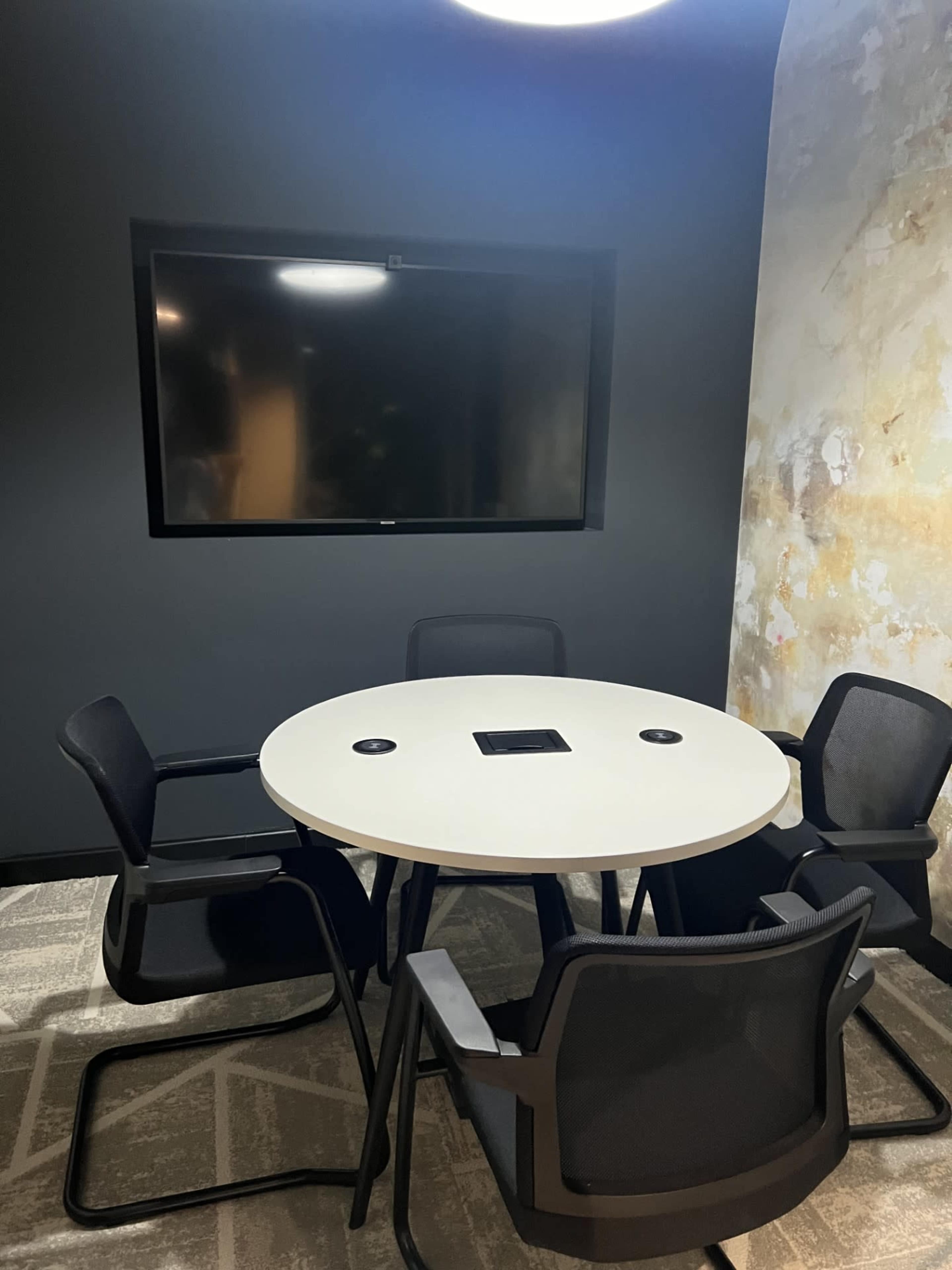 A round table with four chairs is positioned in a small conference room, featuring a wall-mounted television.