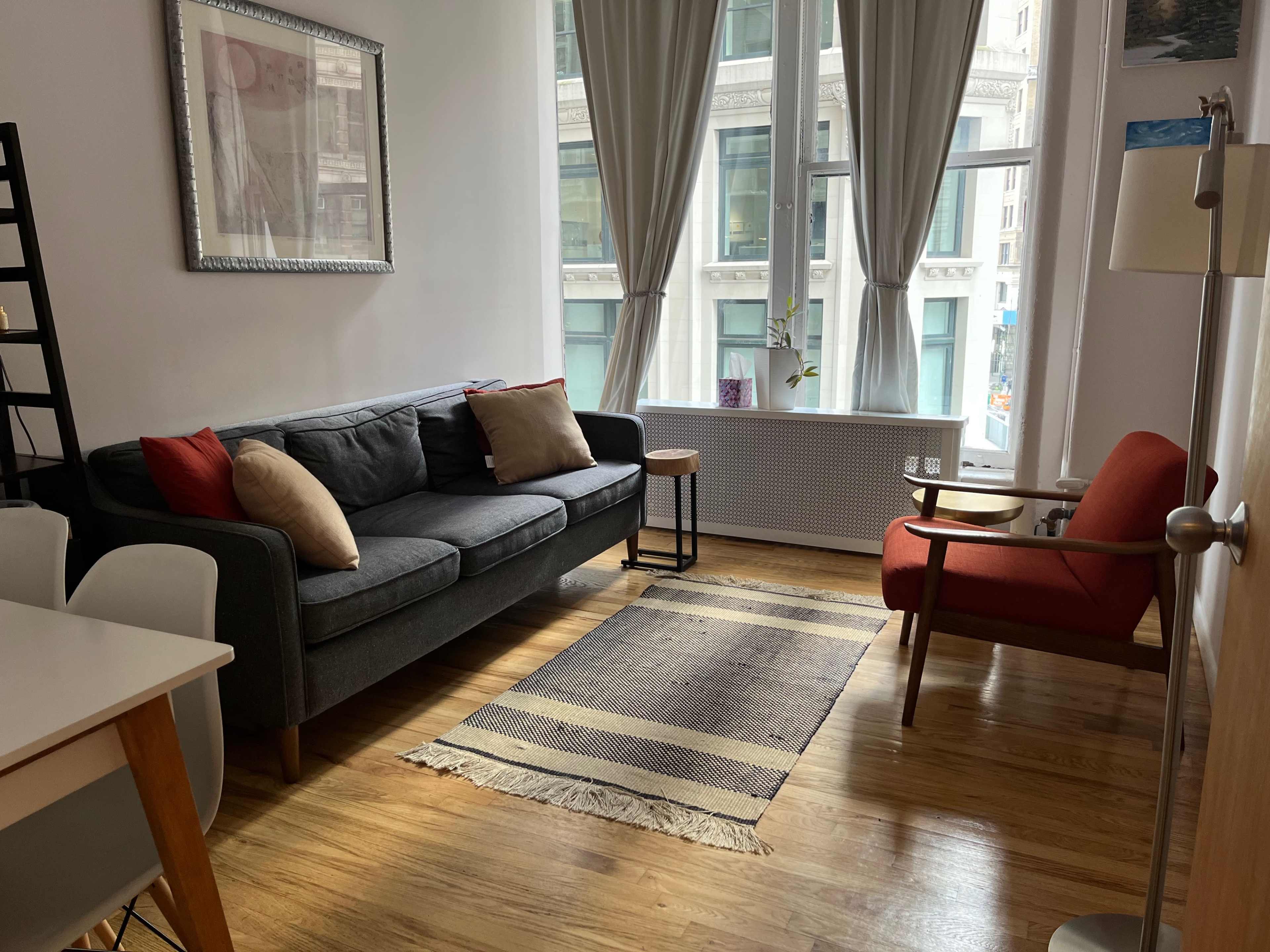 The image shows a cozy living room with a gray sofa, an orange armchair, a striped rug, and large windows allowing natural light to enter.