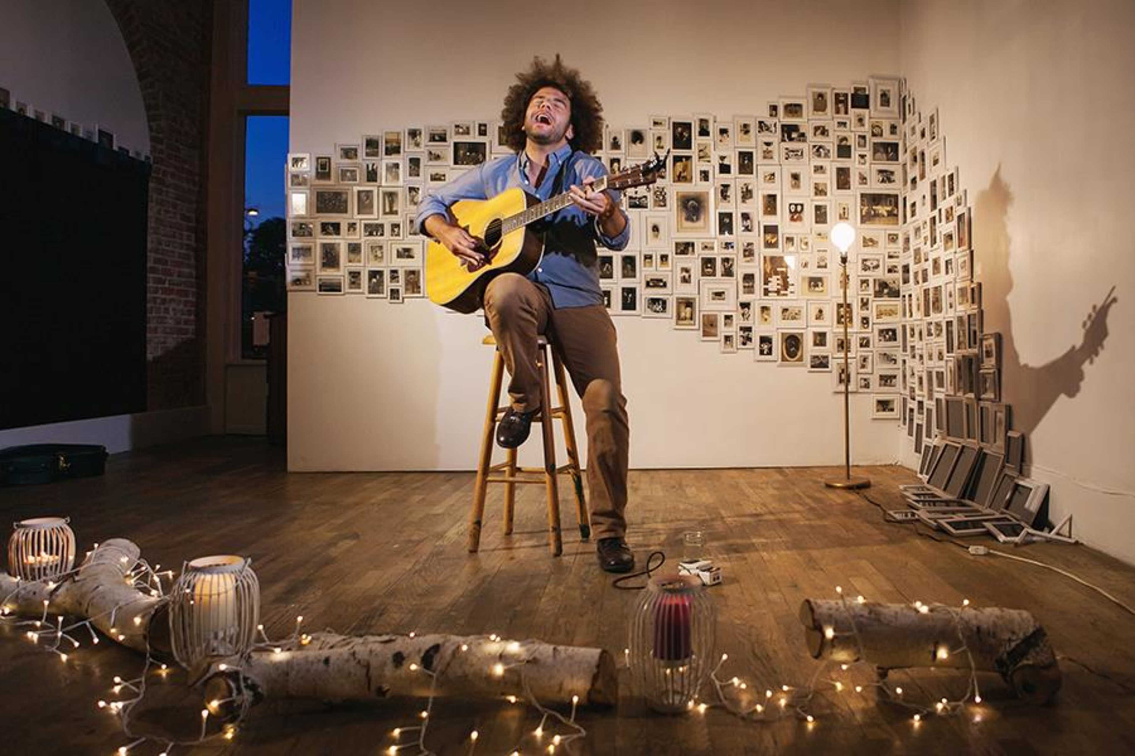 A musician with an afro plays guitar while singing on a stool in a room adorned with numerous framed photographs on the wall.