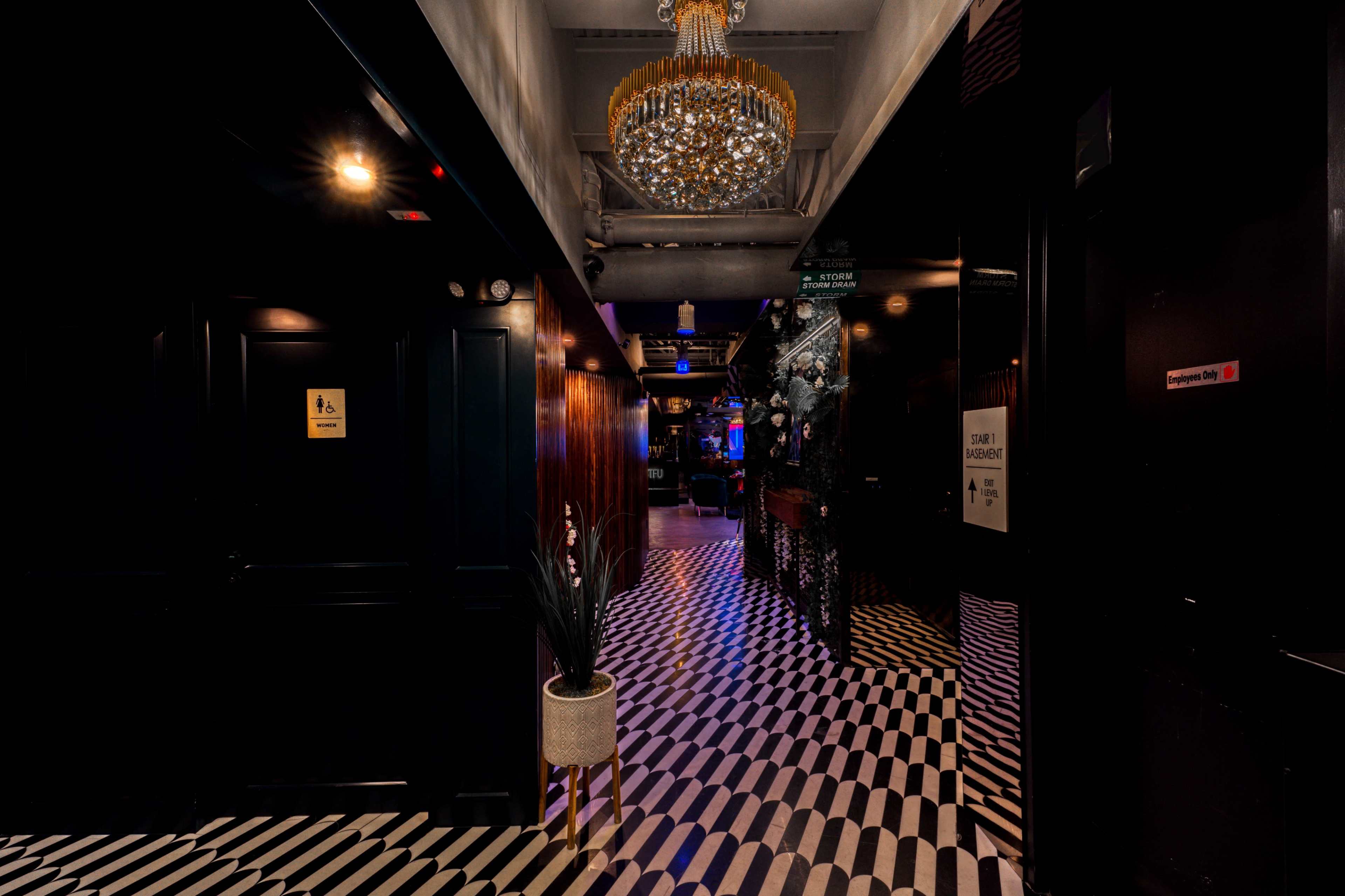 A long hallway with black and white striped flooring, adorned with a chandelier and decorative plants, leads to vibrant lighting at the end.