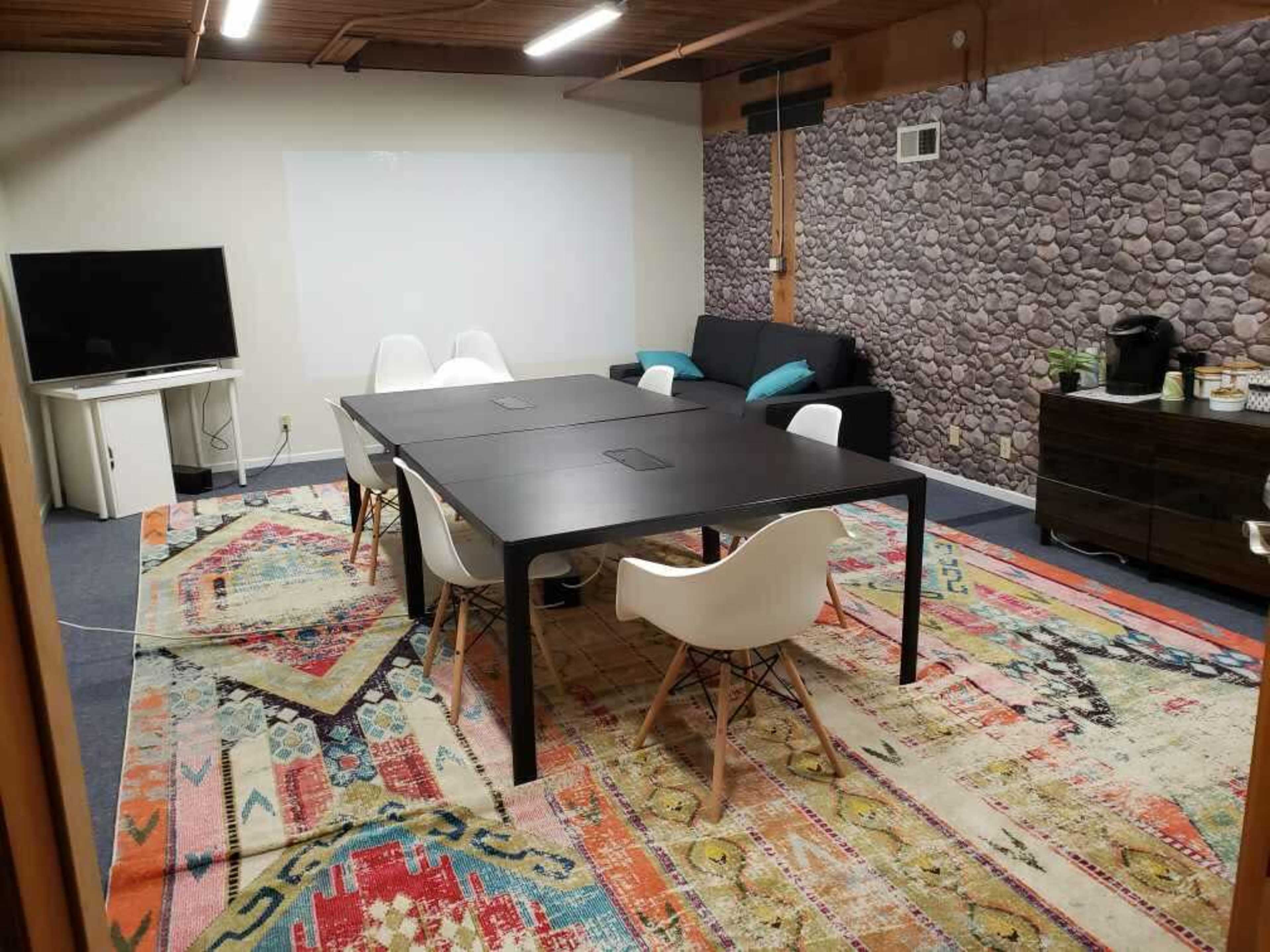 A meeting room features a black table surrounded by white chairs, a television on a stand, a dark sofa, and a patterned rug on the floor.